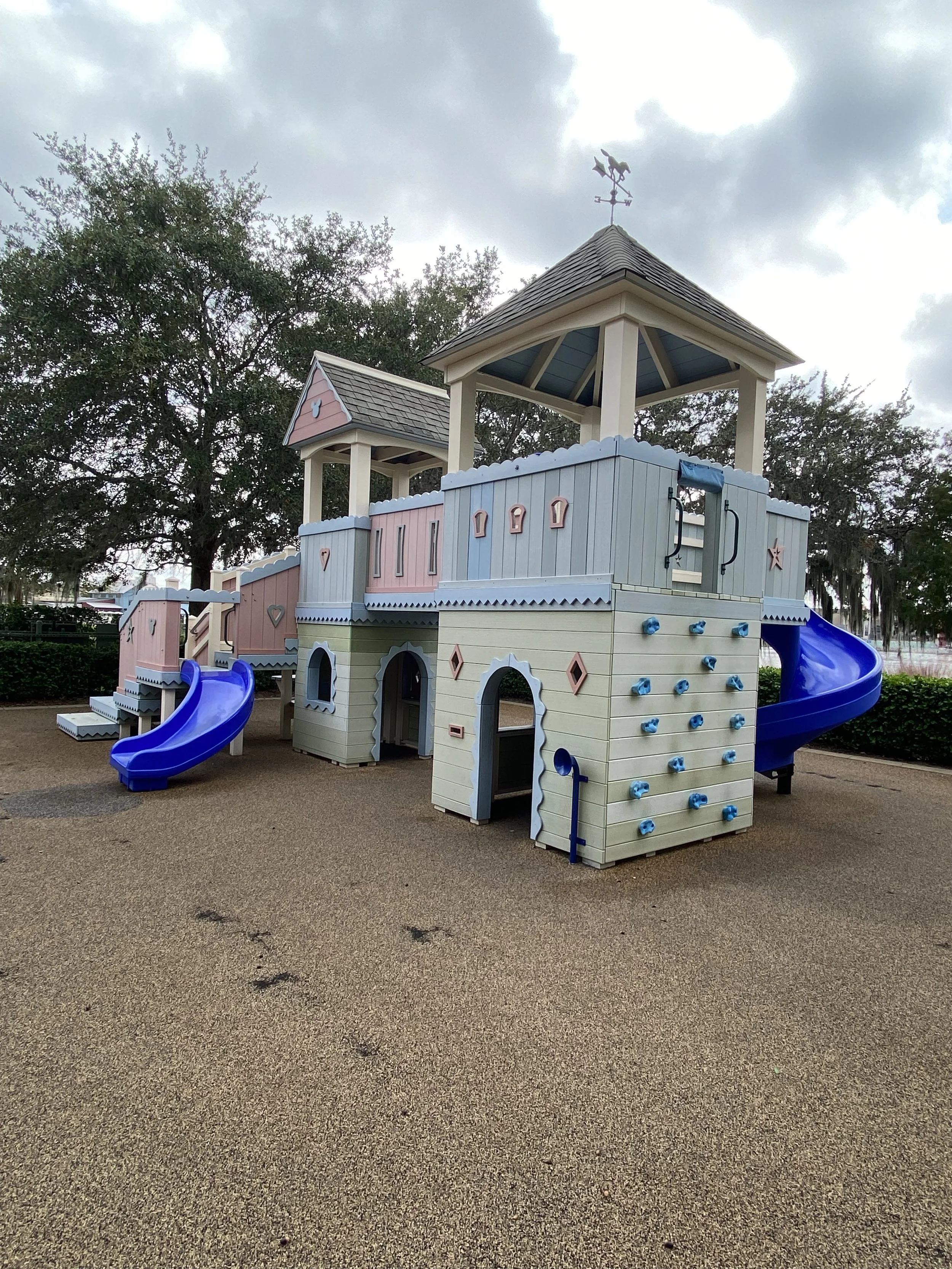 A colorful outdoor playground structure resembling a castle with blue and pink accents, featuring slides, climbing walls, and towers, situated on a gravel surface under cloudy skies with trees in the background.