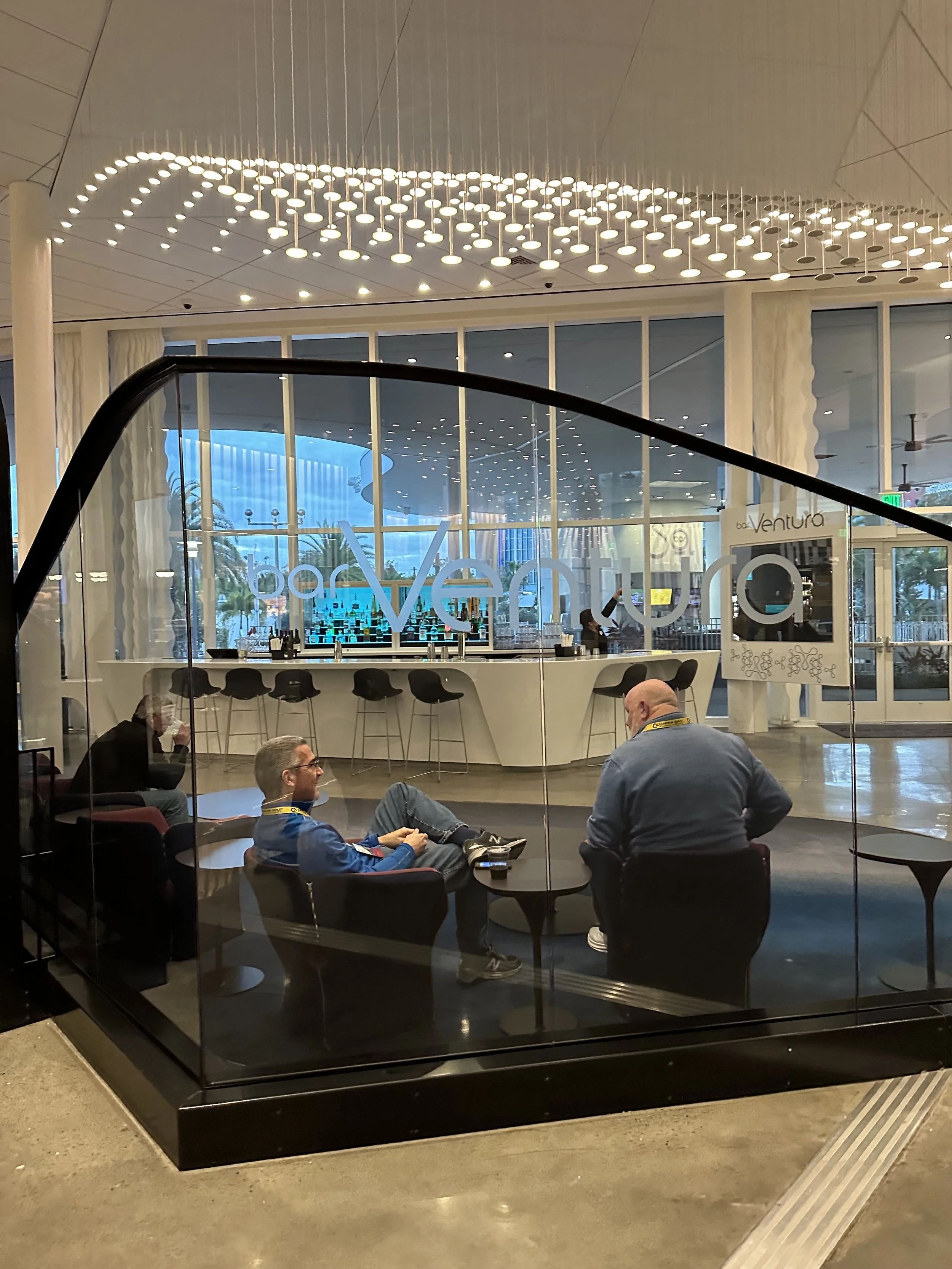People sitting inside a modern, glass-enclosed lounge area at the casino, with a bar counter in the background and decorative ceiling lights.