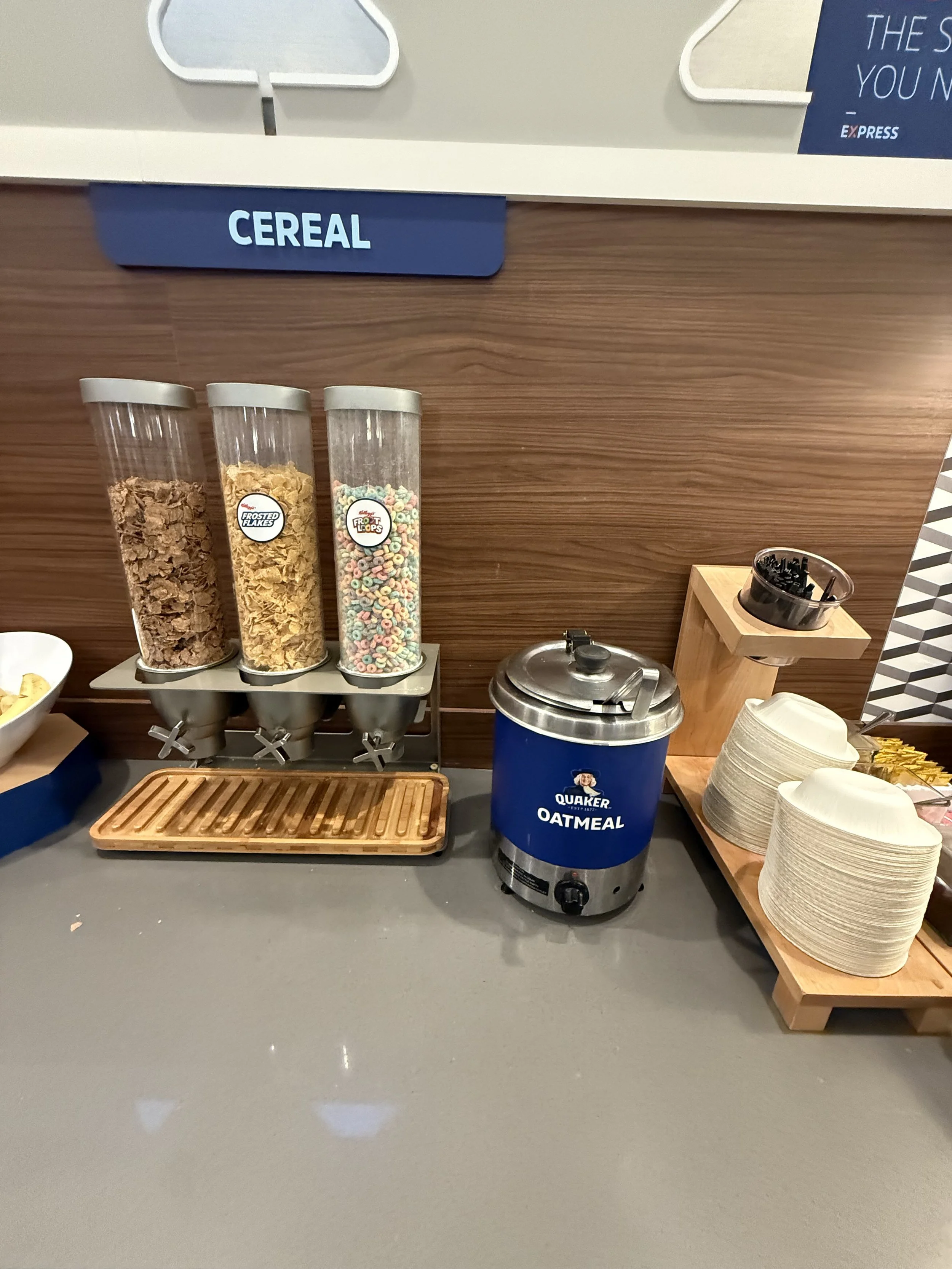 Cereal dispensers with Frosted Flakes, Corn Flakes, and Froot Loops, a container of black spoons, a hot oatmeal dispenser, and stacks of paper bowls.