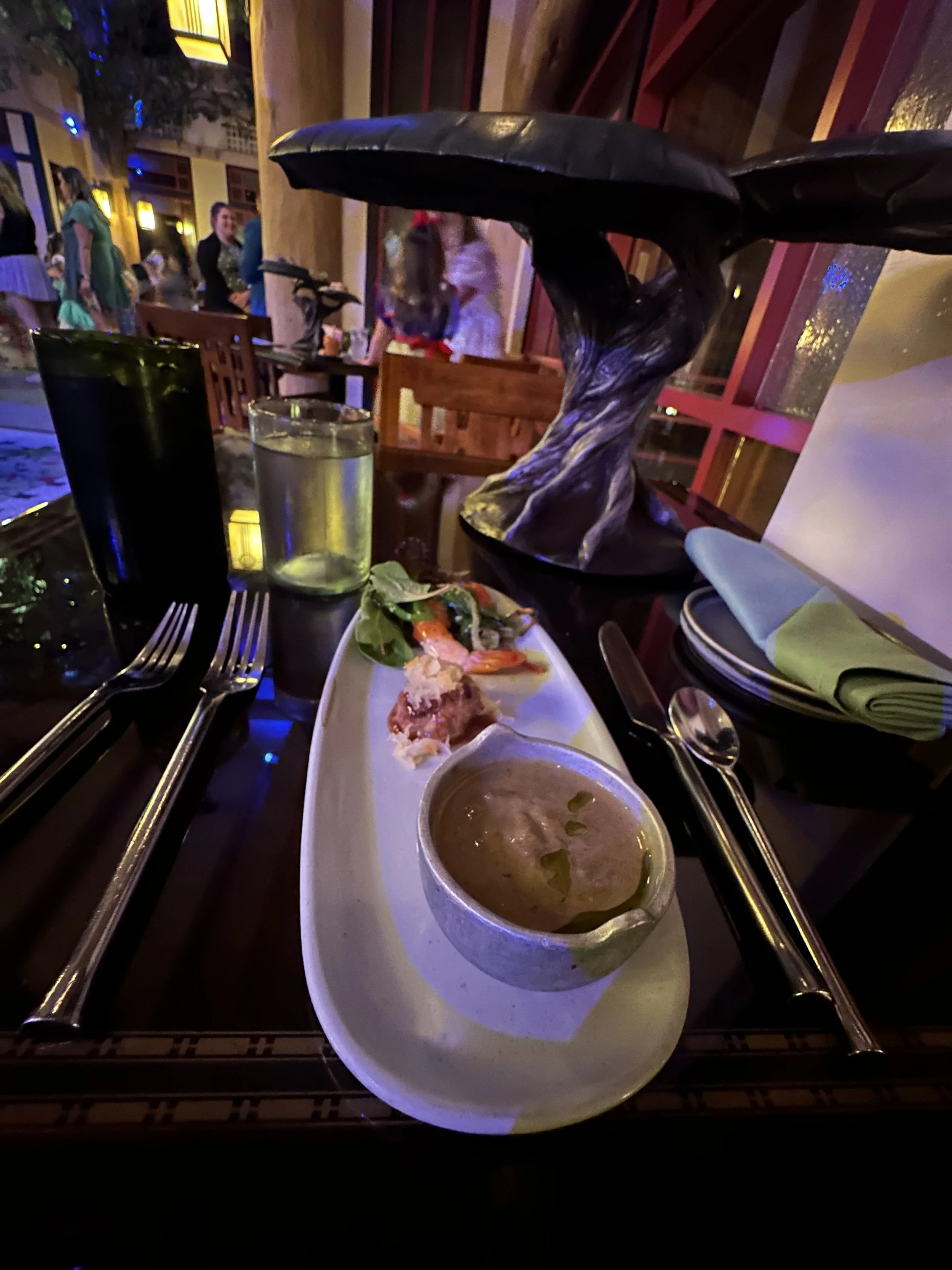 A restaurant table with a bowl of soup, a plate with salad and seafood, a glass of water, a pitcher, and utensils, with a decorative sculpture and other diners in the background.