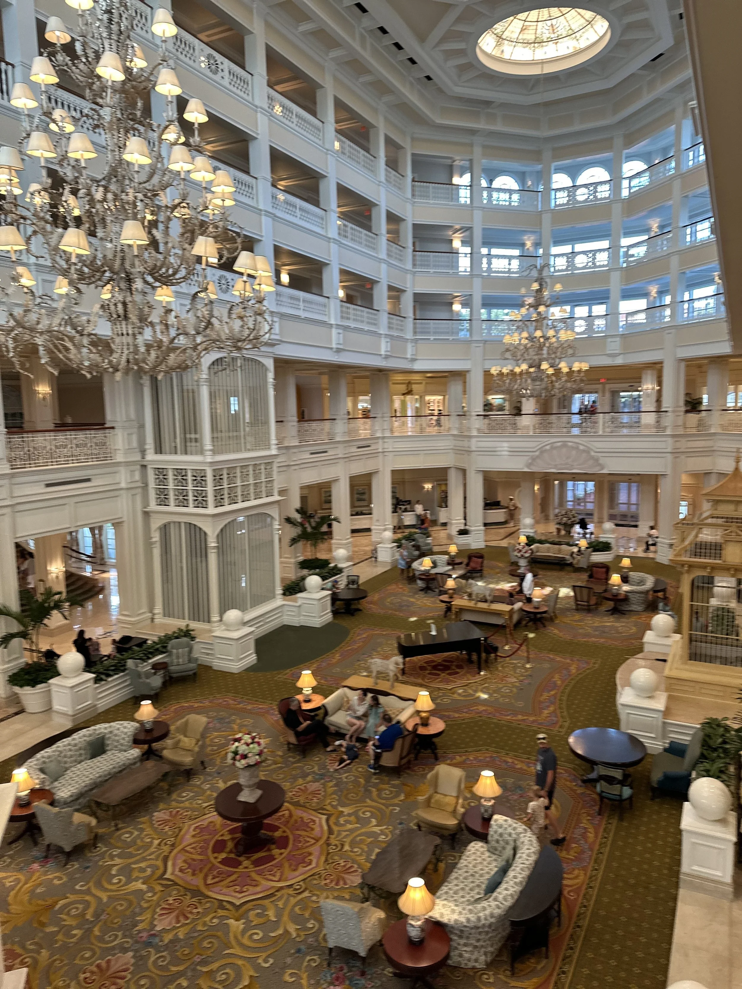 Interior of a luxurious hotel lobby with high ceilings, large chandeliers, multiple floors, and elegant seating areas with people relaxing.