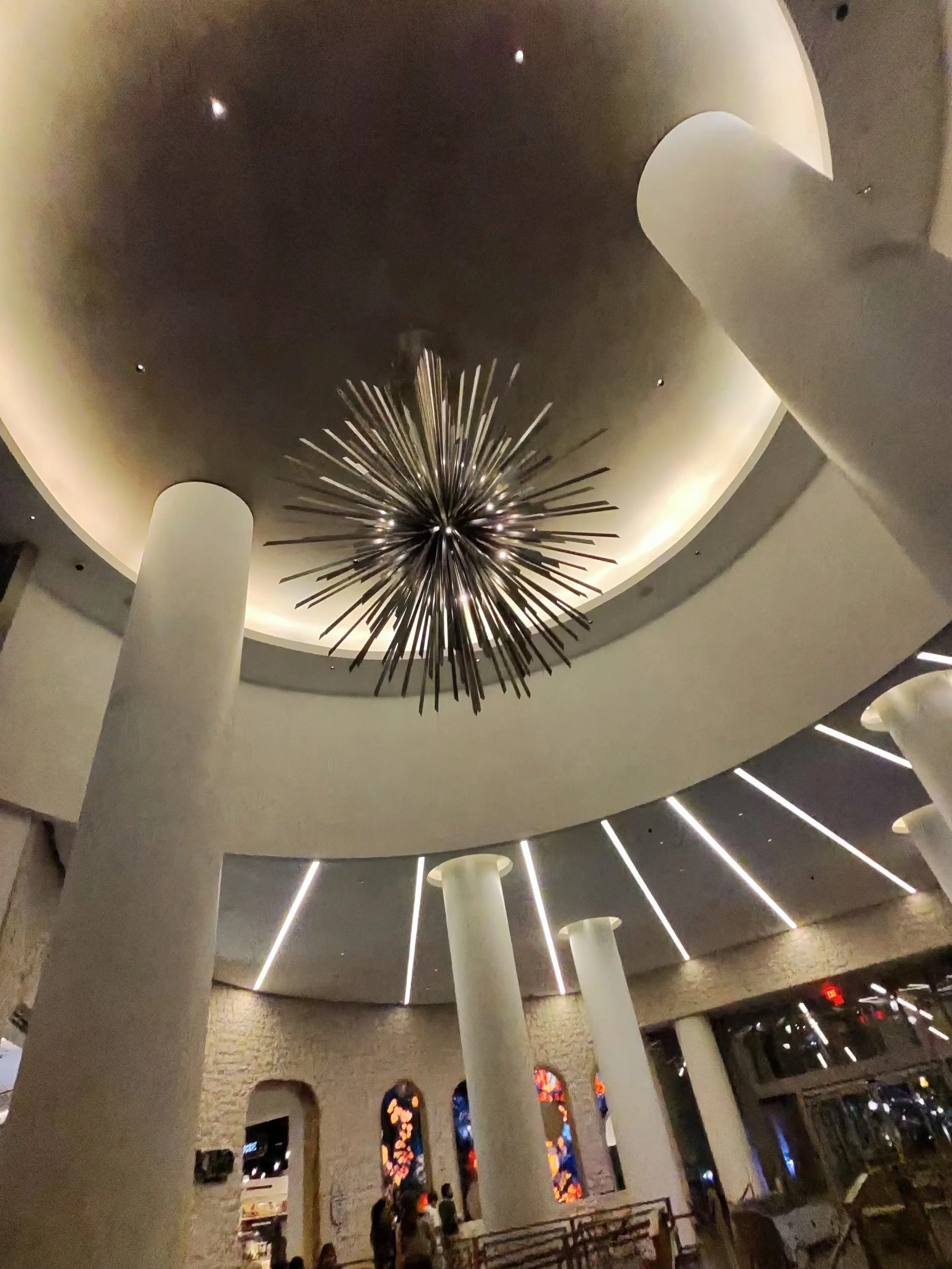 A modern interior with a circular ceiling featuring a large, starburst chandelier hanging from the center. The ceiling has recessed lighting along the circular edge, and tall white columns support the structure. Stained glass windows and people near 