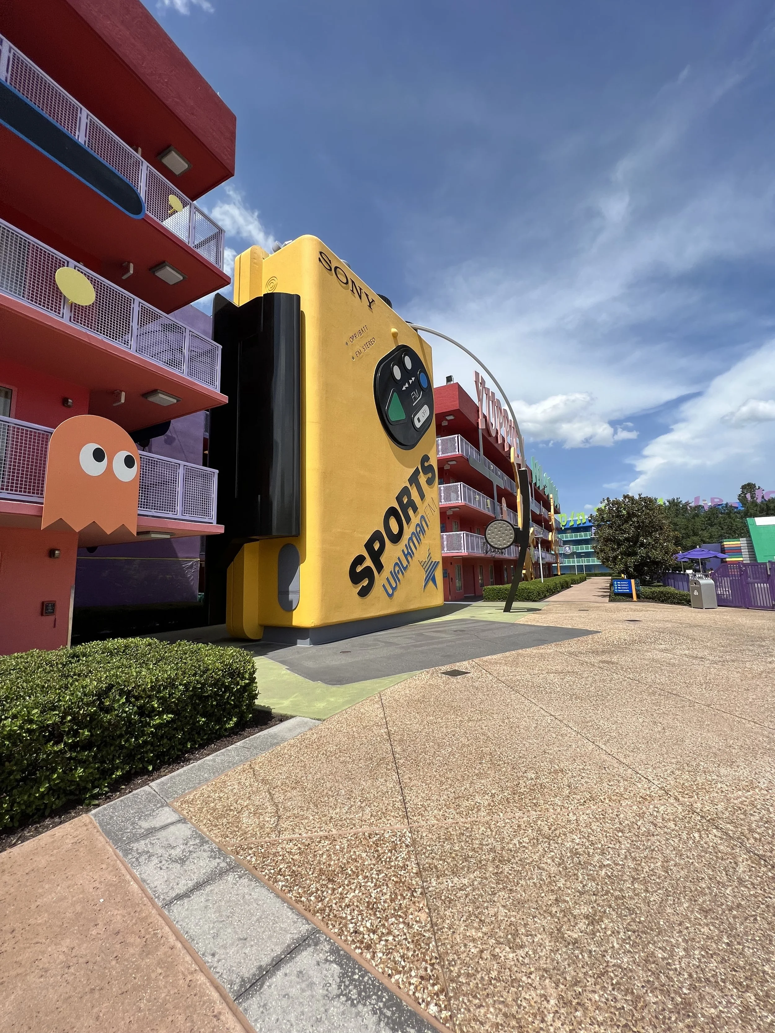 Large yellow sign shaped like a camera with 'SPORTS' written on it, located outside a colorful themed building with purple, pink, and blue colors and a cartoon ghost figure on the left side, under a bright blue sky at an amusement park or similar att