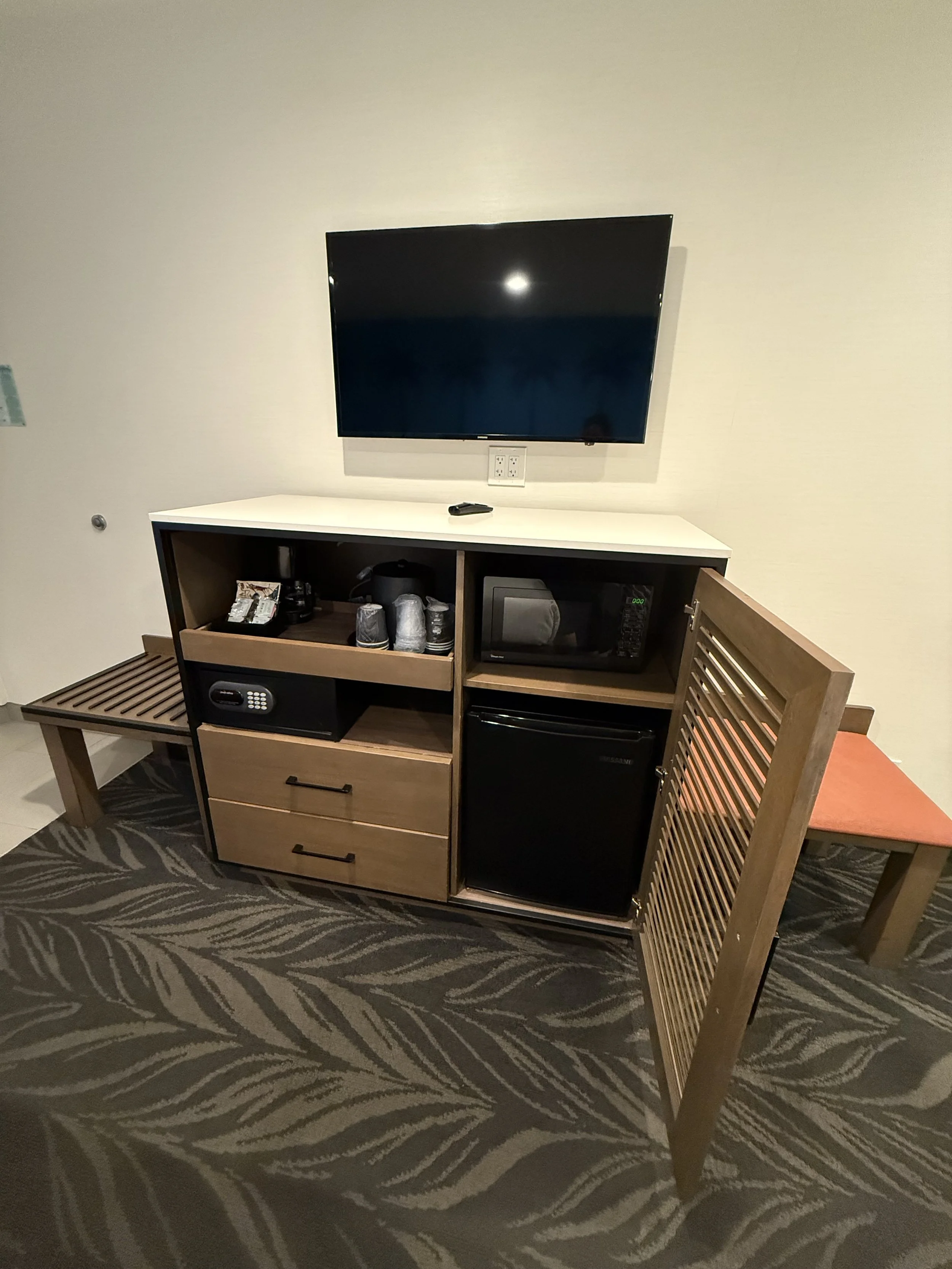 A hotel room minibar with a small refrigerator, microwave, safe, and coffee supplies, beside a small bench, and a wall-mounted flat-screen TV above.