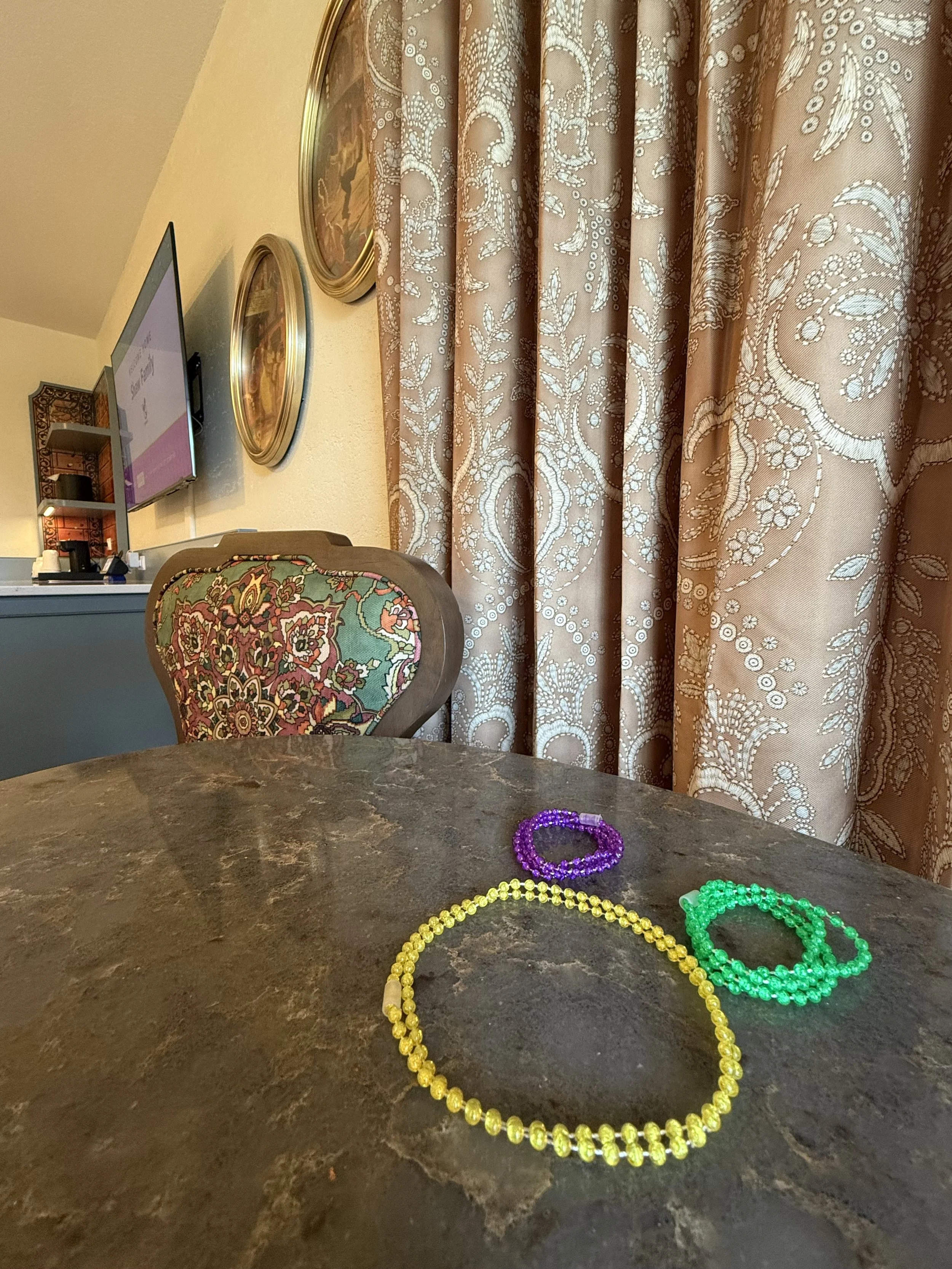 Colorful beaded necklaces, including purple, yellow, and green, are on a dark brown marble table in a room with patterned curtains and vintage-style wall decor.