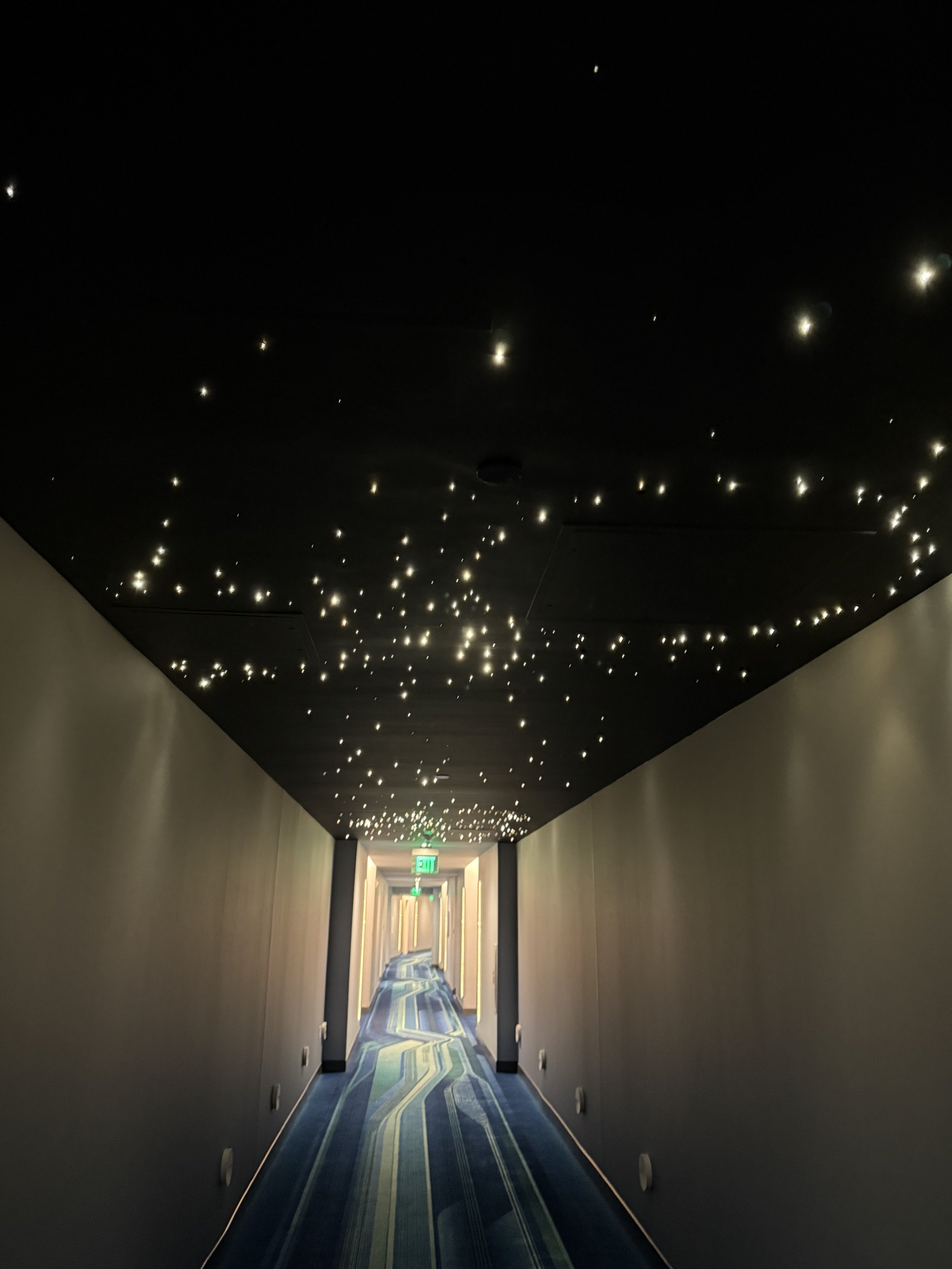Long hotel hallway with dark ceiling decorated with small star-shaped LED lights, patterned blue and green carpet, and an illuminated green exit sign at the end.
