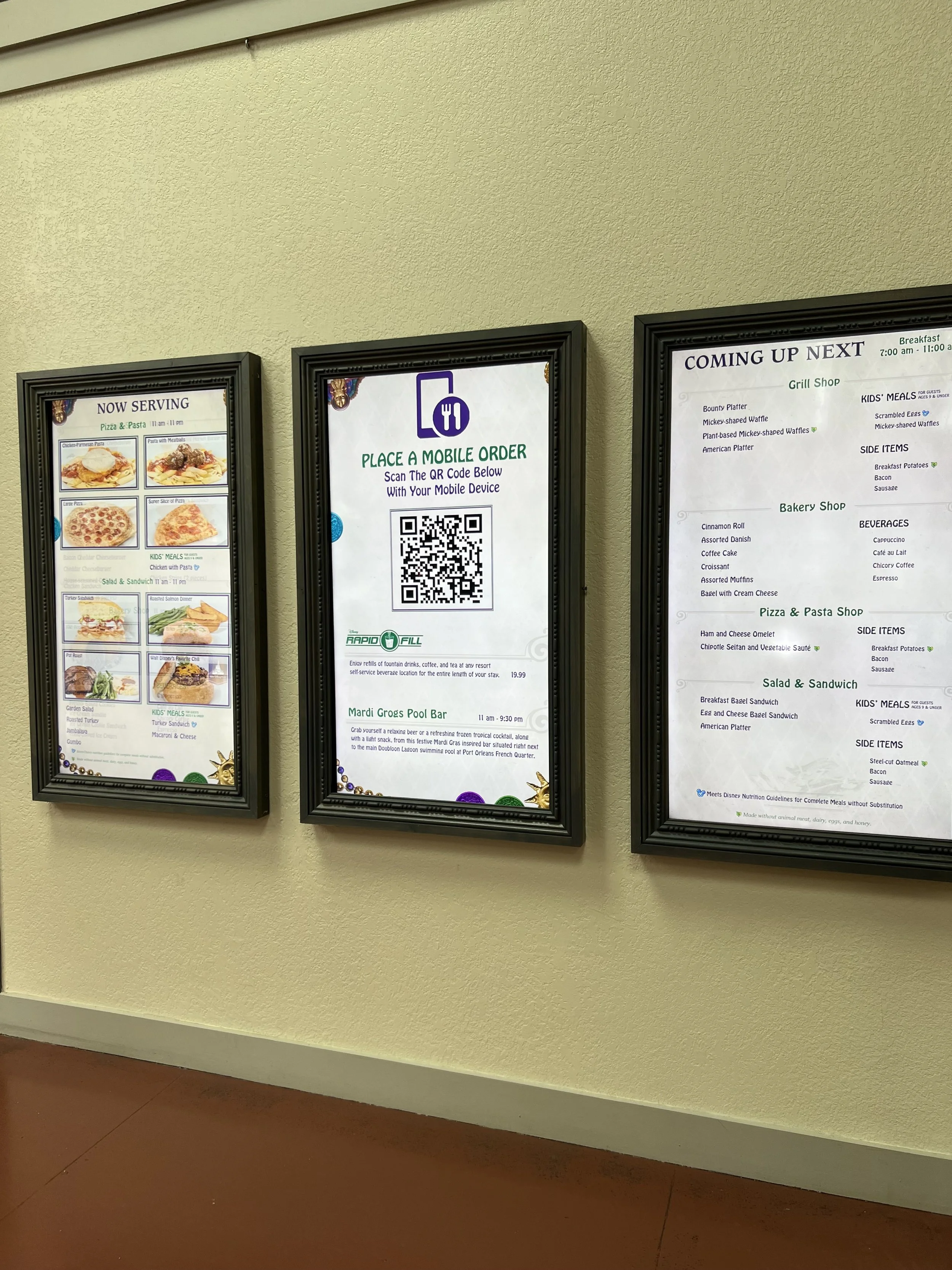 Three framed menu boards on a yellow wall showing food options, QR code for mobile orders, and upcoming meal schedules.