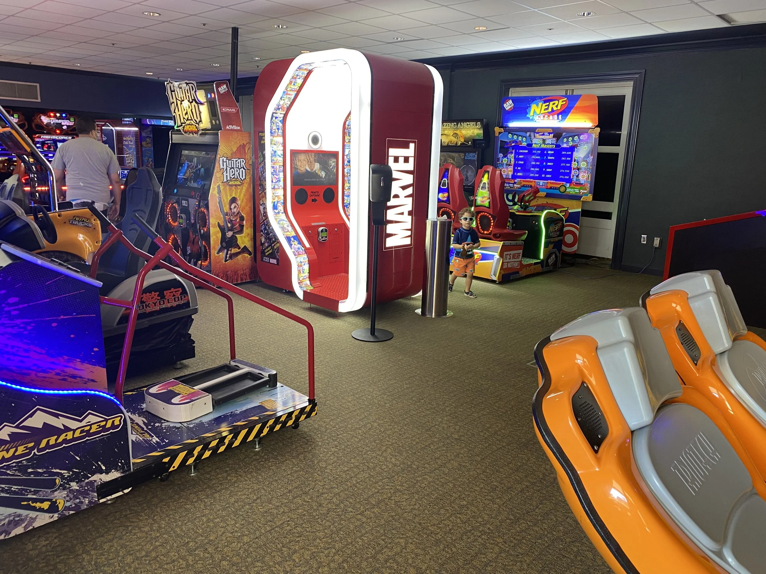 Indoor arcade with various gaming machines, including a Marvel-themed game, a Nerf arcade, racing seats, and sound-based games, with a young boy wearing sunglasses standing near the arcade machines.