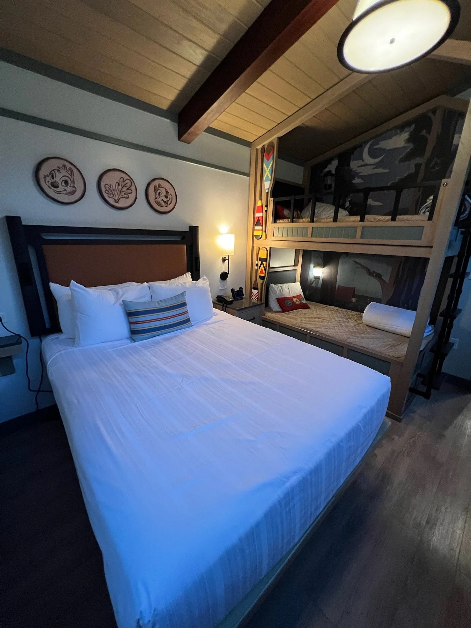 A hotel room with a large bed dressed in white linens and four pillows, a small striped decorative pillow, wall decor featuring cartoon animal faces, a bedside lamp, a bunk bed with a natural wood frame, and a dark mural of a night sky with a moon an