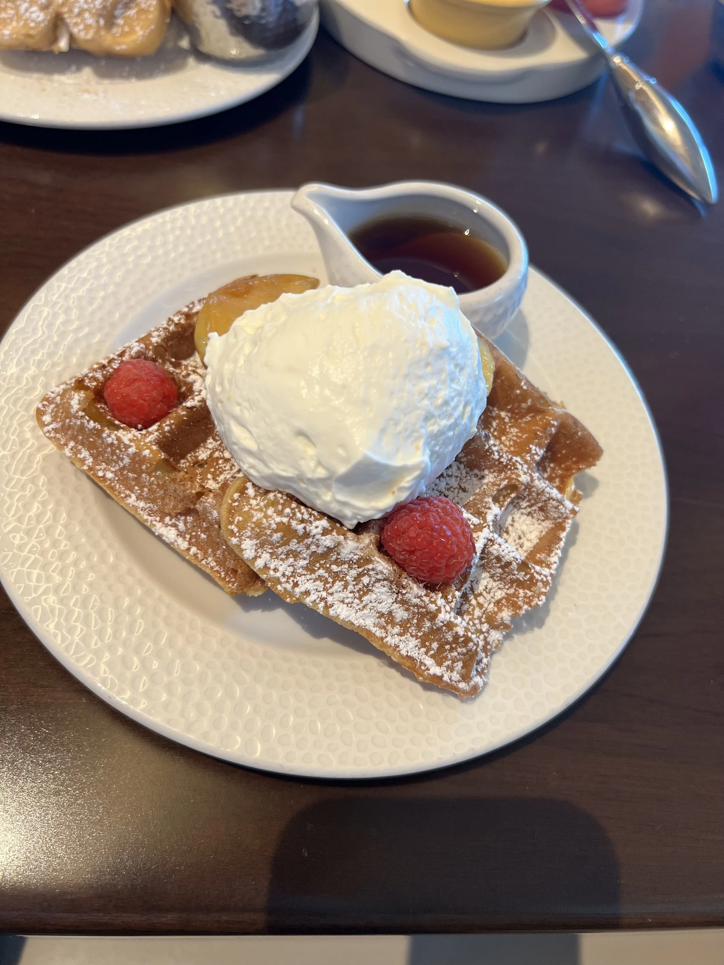 A plate with Belgian waffle topped with whipped cream, raspberries, and powdered sugar, served with a side of syrup.