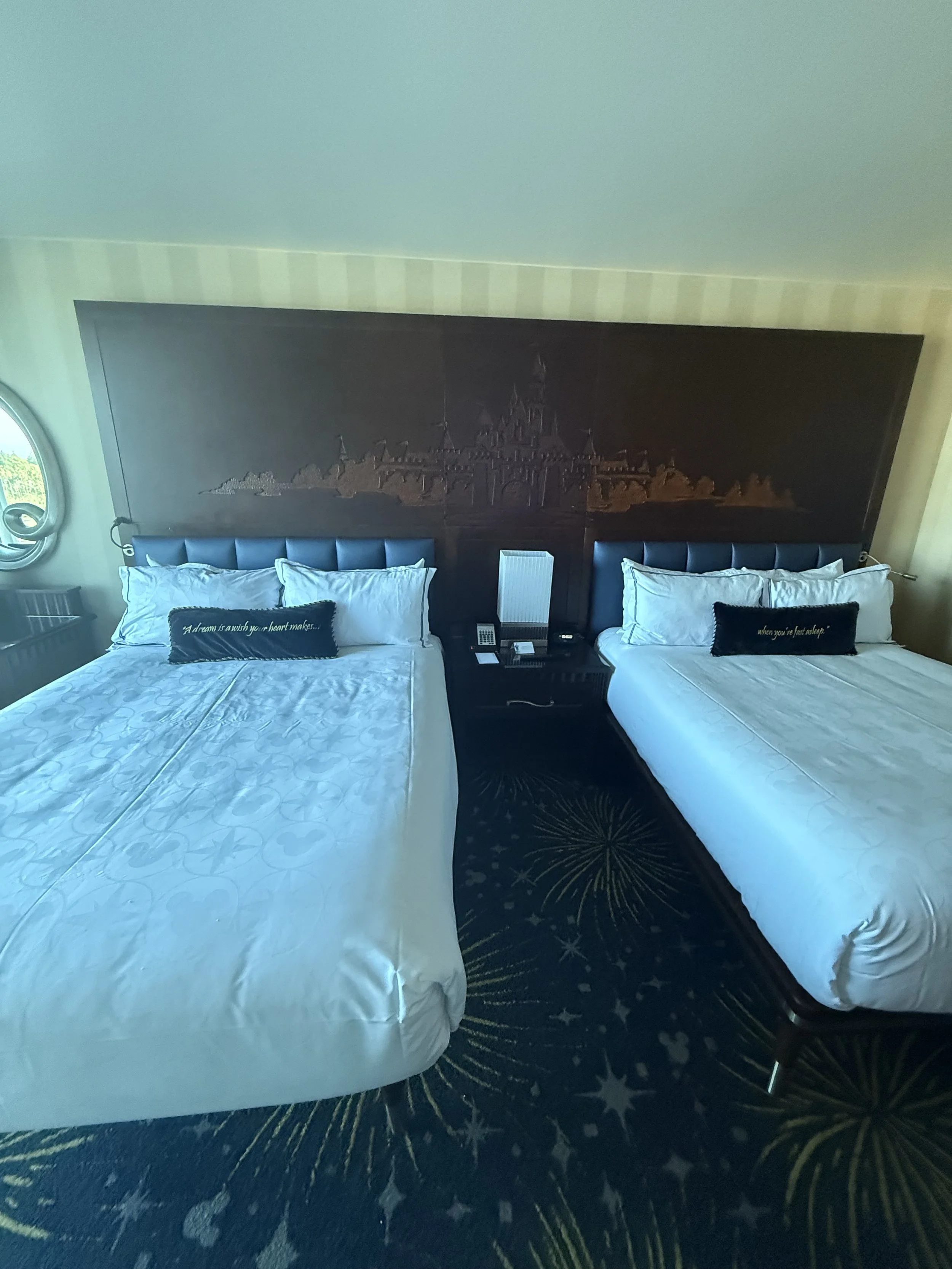 Two beds with white bedding and black headboards in a hotel room, with a dark wooden wall featuring a carved castle scene above the beds, and a nightstand between the beds holding a phone and a lamp. A window with a circular design is on the left sid