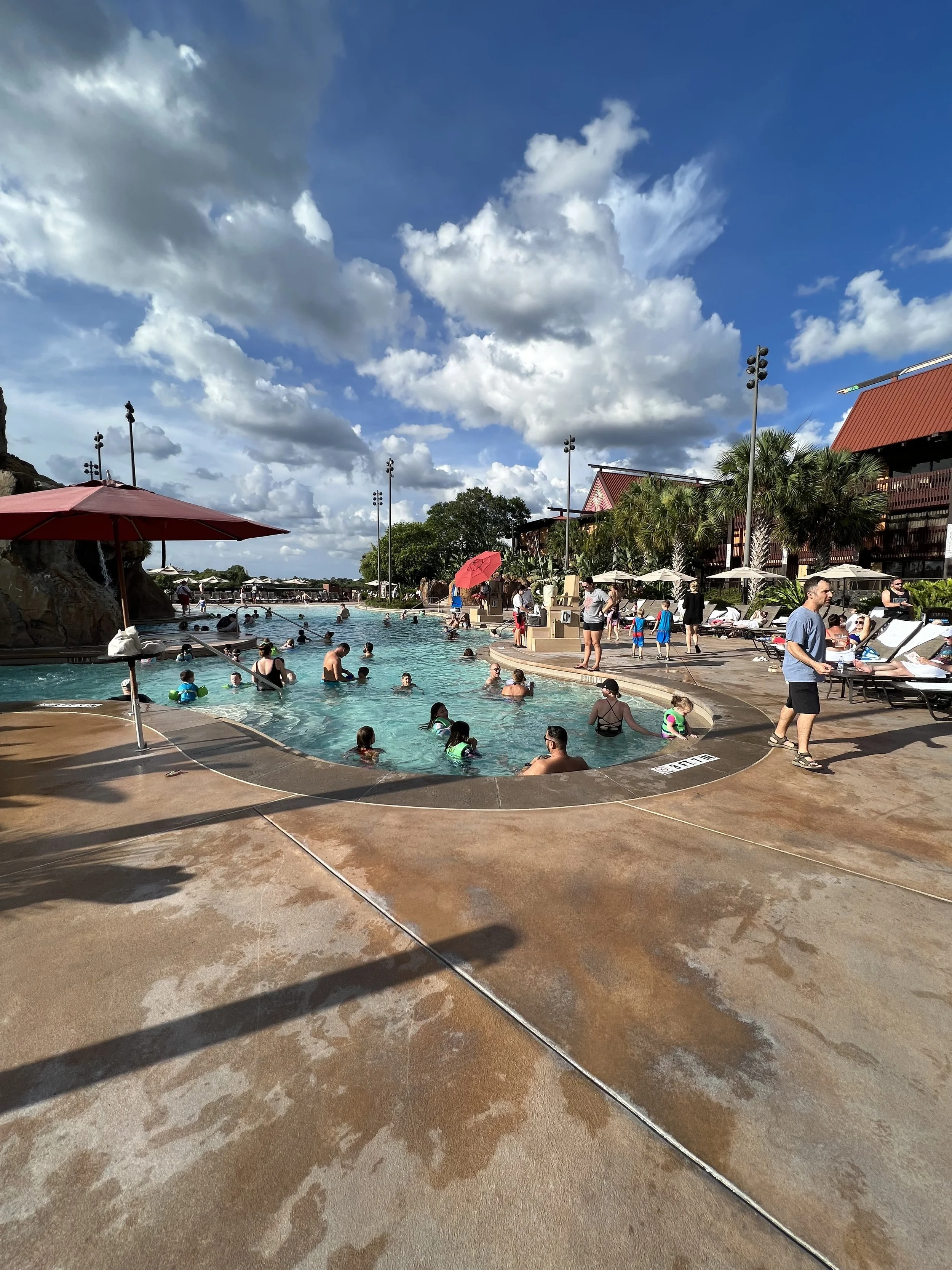 People swimming and relaxing at a large outdoor waterpark with umbrellas, lounge chairs, and palm trees, under a sky with clouds.