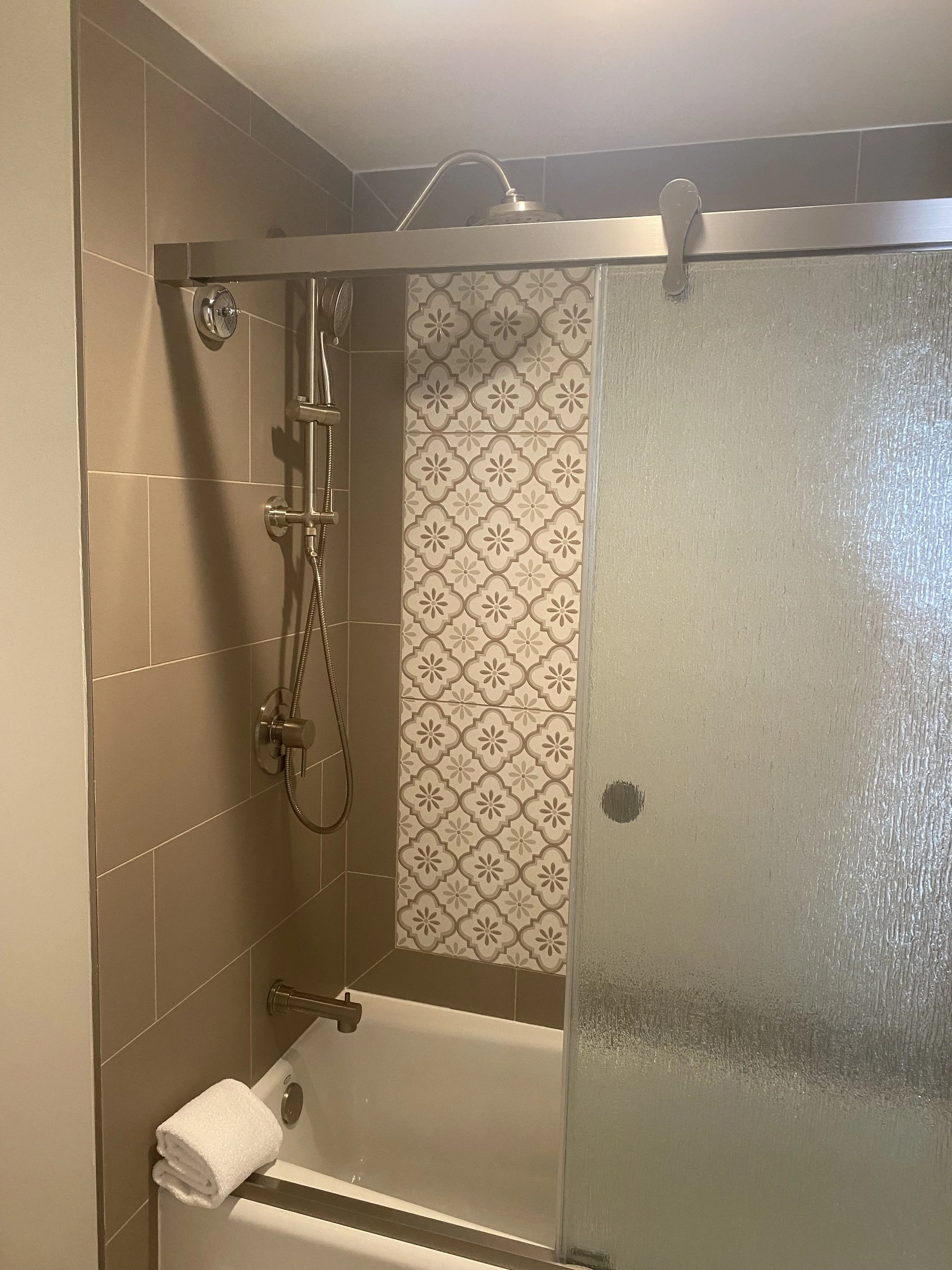 A bathroom shower area with tan and patterned tiles, a sliding textured glass door, and a towel hanging on the bathtub.