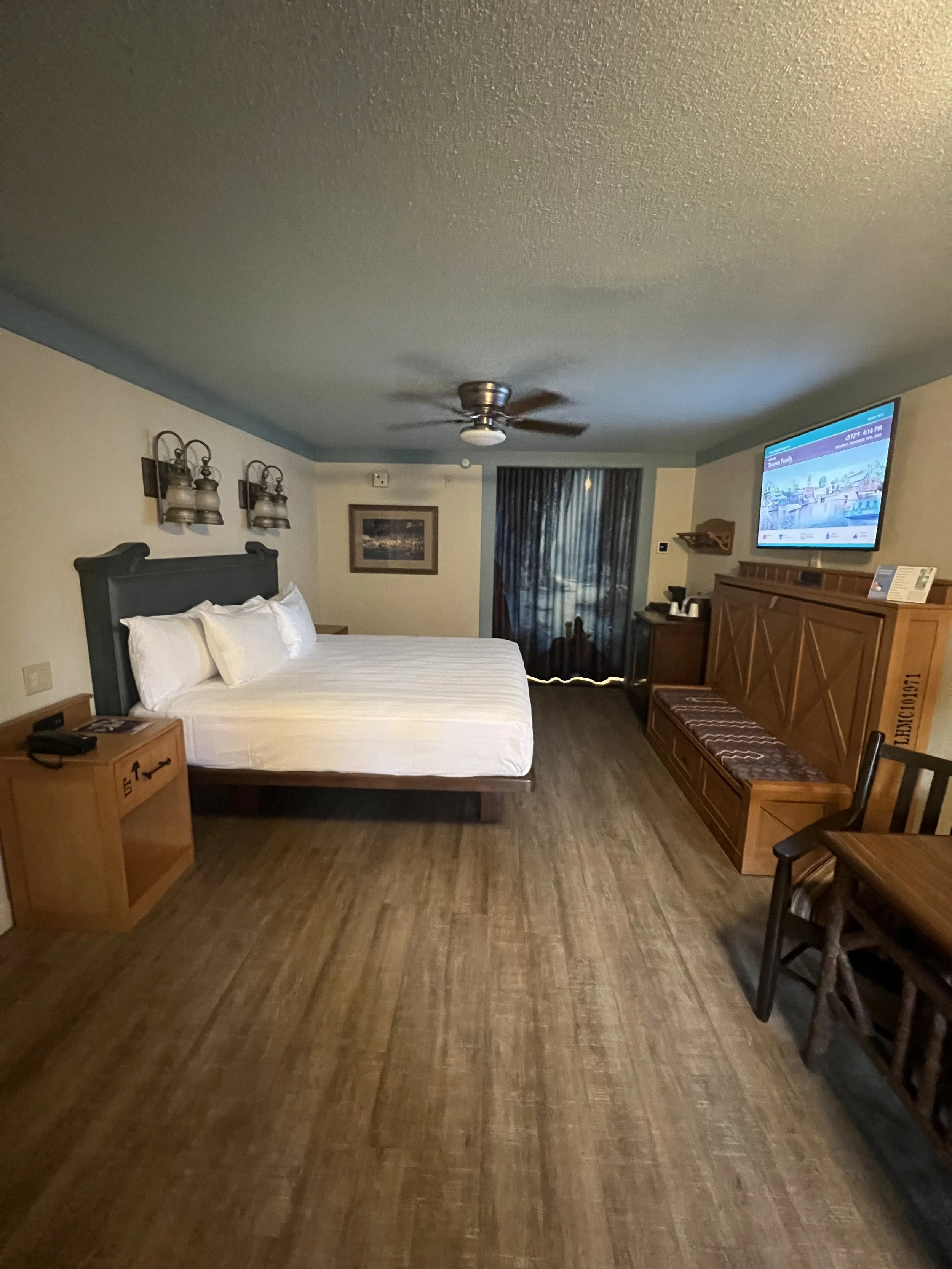 Hotel room featuring a king-size bed with white linens, a wooden nightstand, wall-mounted lamps, a framed picture, a TV mounted on the wall, a wooden bench, a small table, chairs, a curtain, and a ceiling fan.