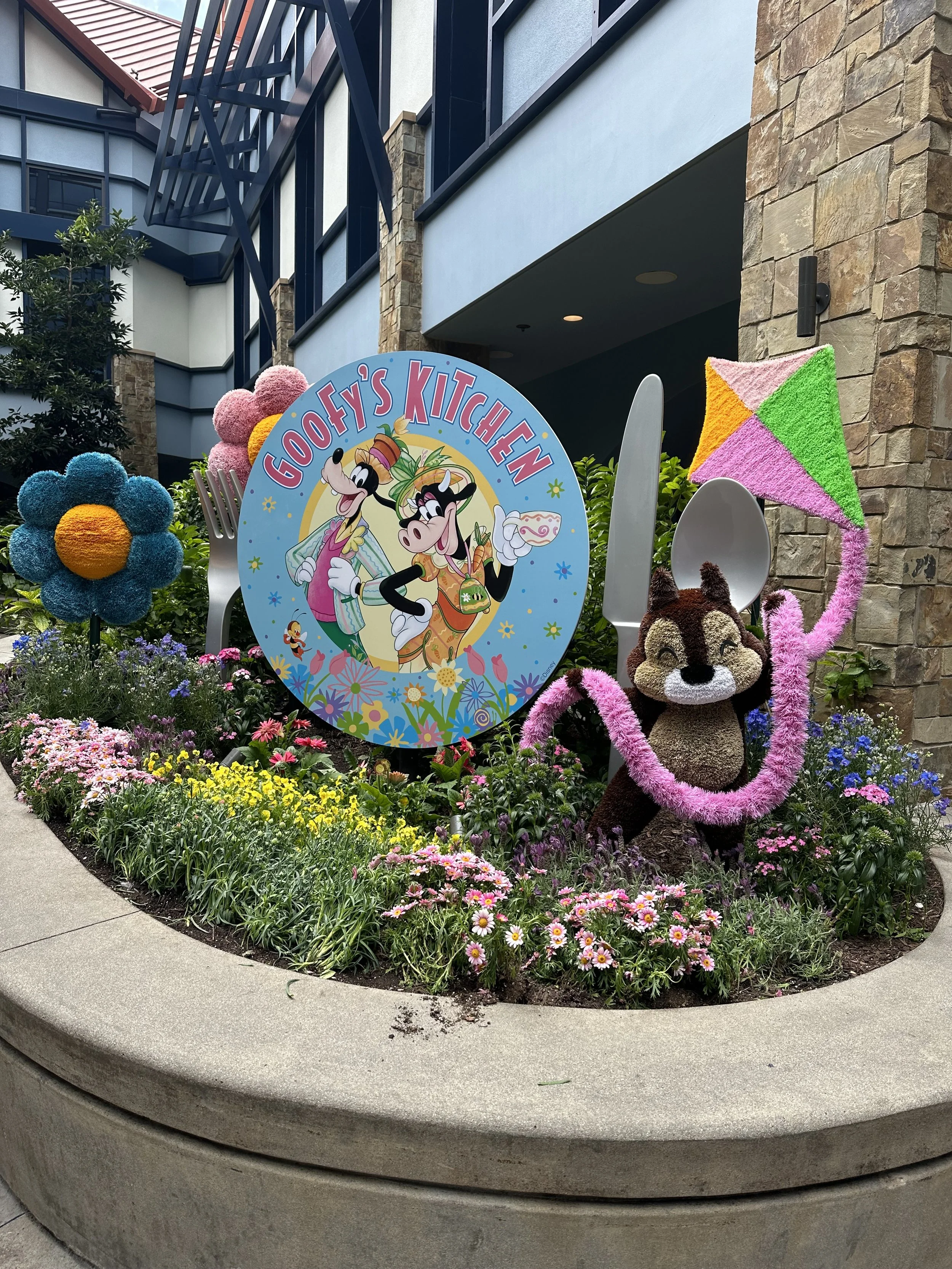 Colorful outdoor display with a sign reading 'Goofy’s Kitchen' featuring animated characters Goofy and Daisy Duck, surrounded by large floral sculptures, a stuffed chipmunk holding a pink kite, and various flowers in a garden bed.