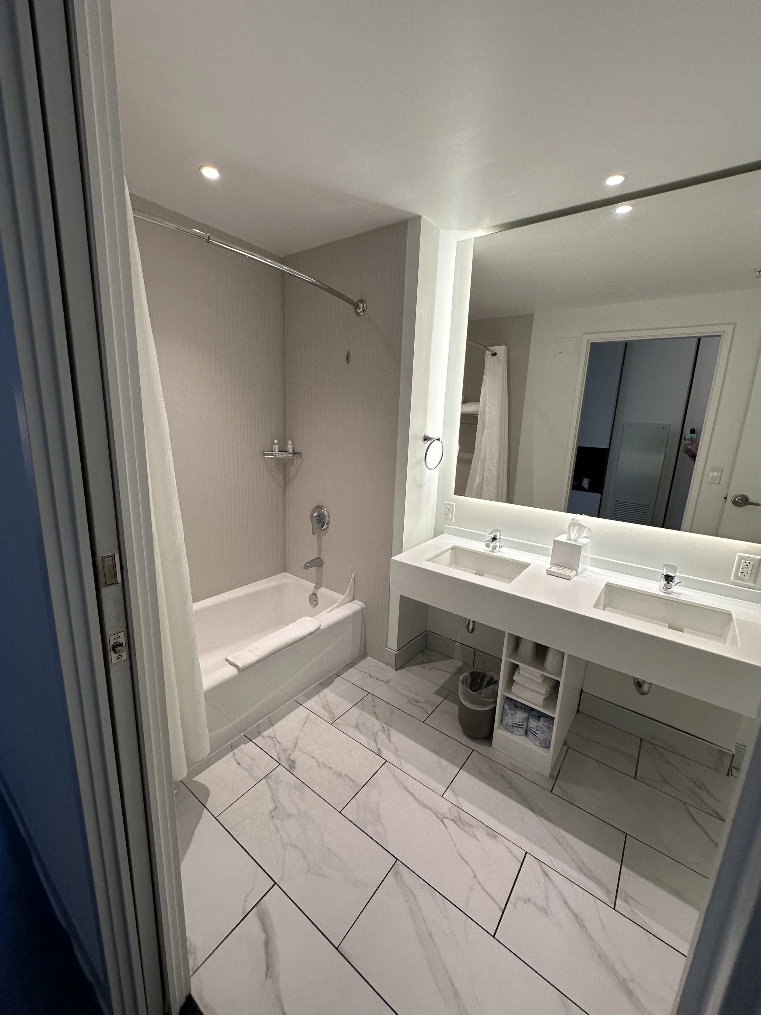 Modern bathroom with a bathtub, shower curtain, dual sinks, large mirror, and white marble floor tiles.