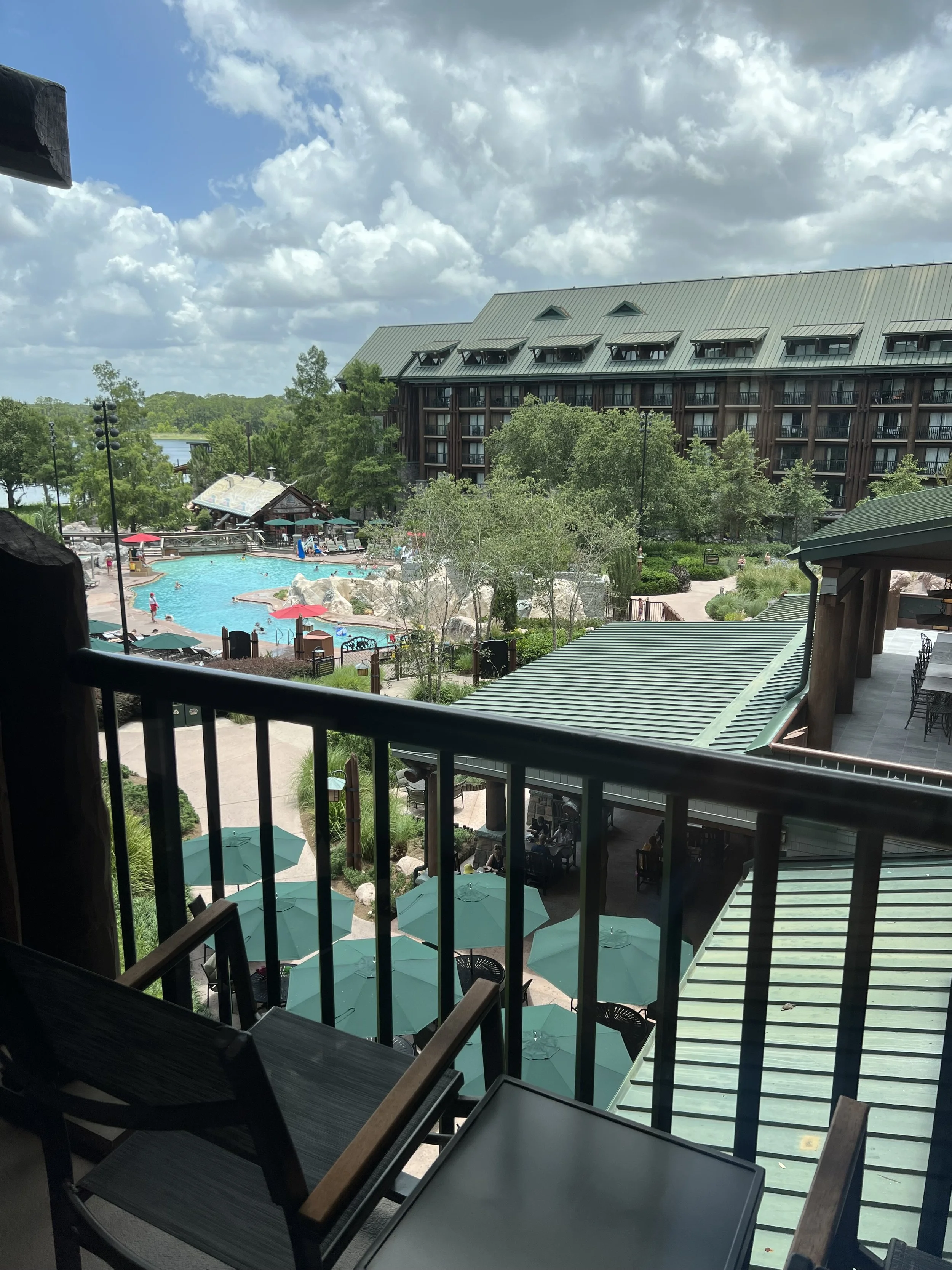 View from a balcony overlooking a resort pool area with umbrellas, lounge chairs, and surrounding trees, with a large multi-story building in the background under a partly cloudy sky.