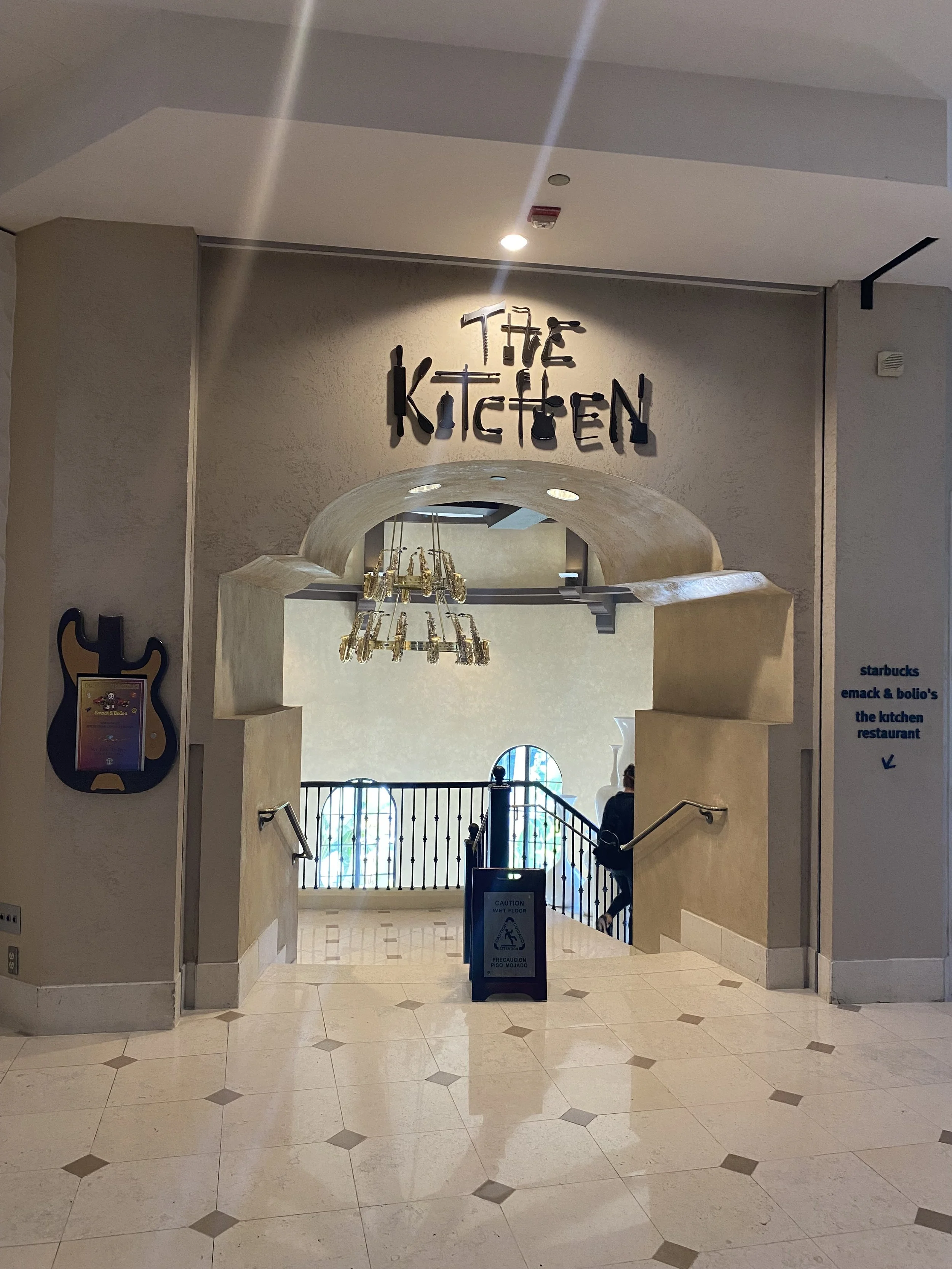 Entrance to a restaurant called 'The Kitchen,' with stairs leading down, a chandelier hanging, and signage for Starbucks and other dining options on the side wall.