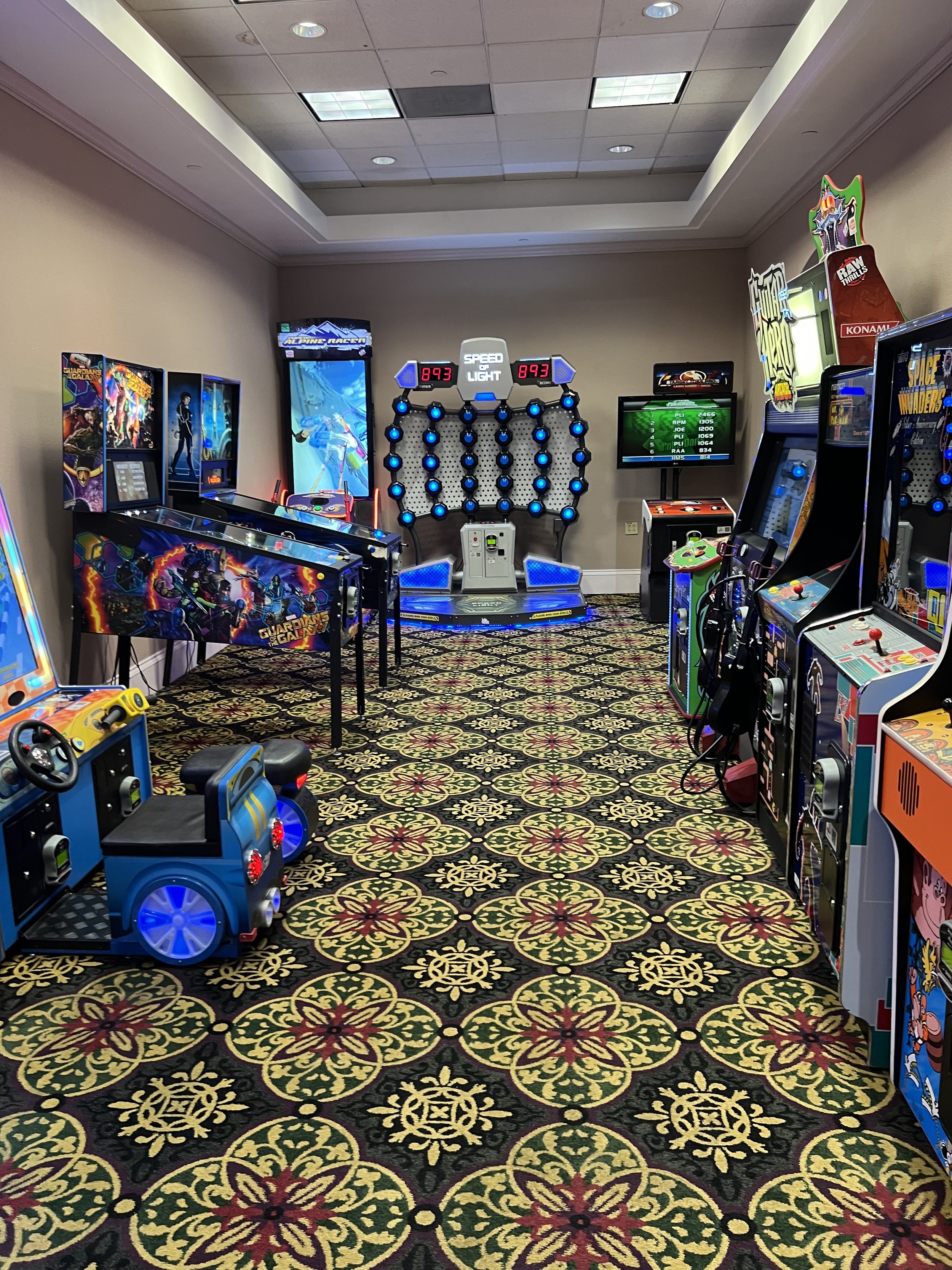 Arcade room with various vintage arcade games, a blue car ride game on the left, and a dance game in the center with blue lights and a digital display. The room has patterned carpet and beige walls.