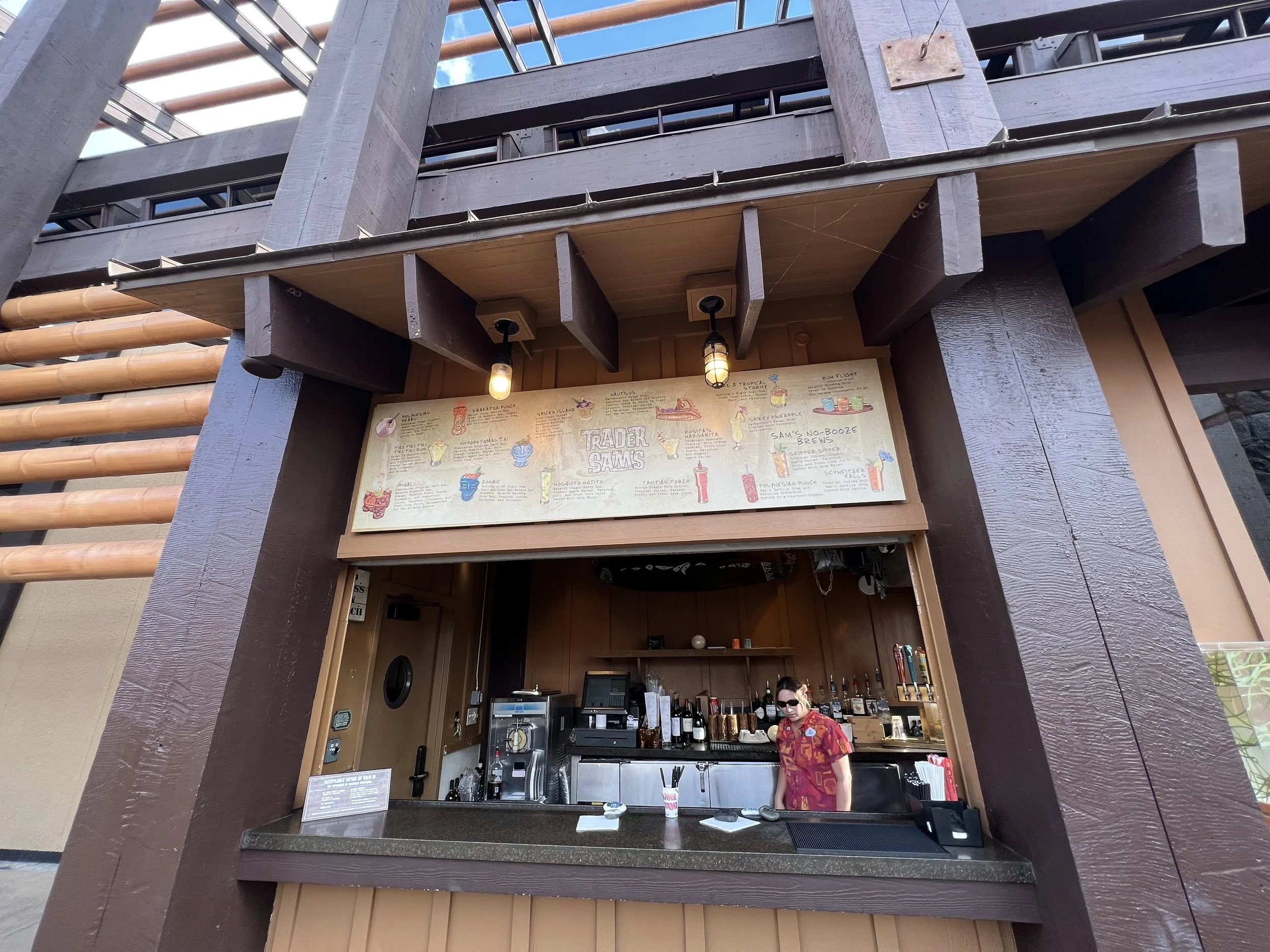 A bar or drink stand with a person wearing sunglasses and a red Hawaiian shirt, behind the bar counter with various bottles and drink equipment, under a sign with illustrated drink recipes.