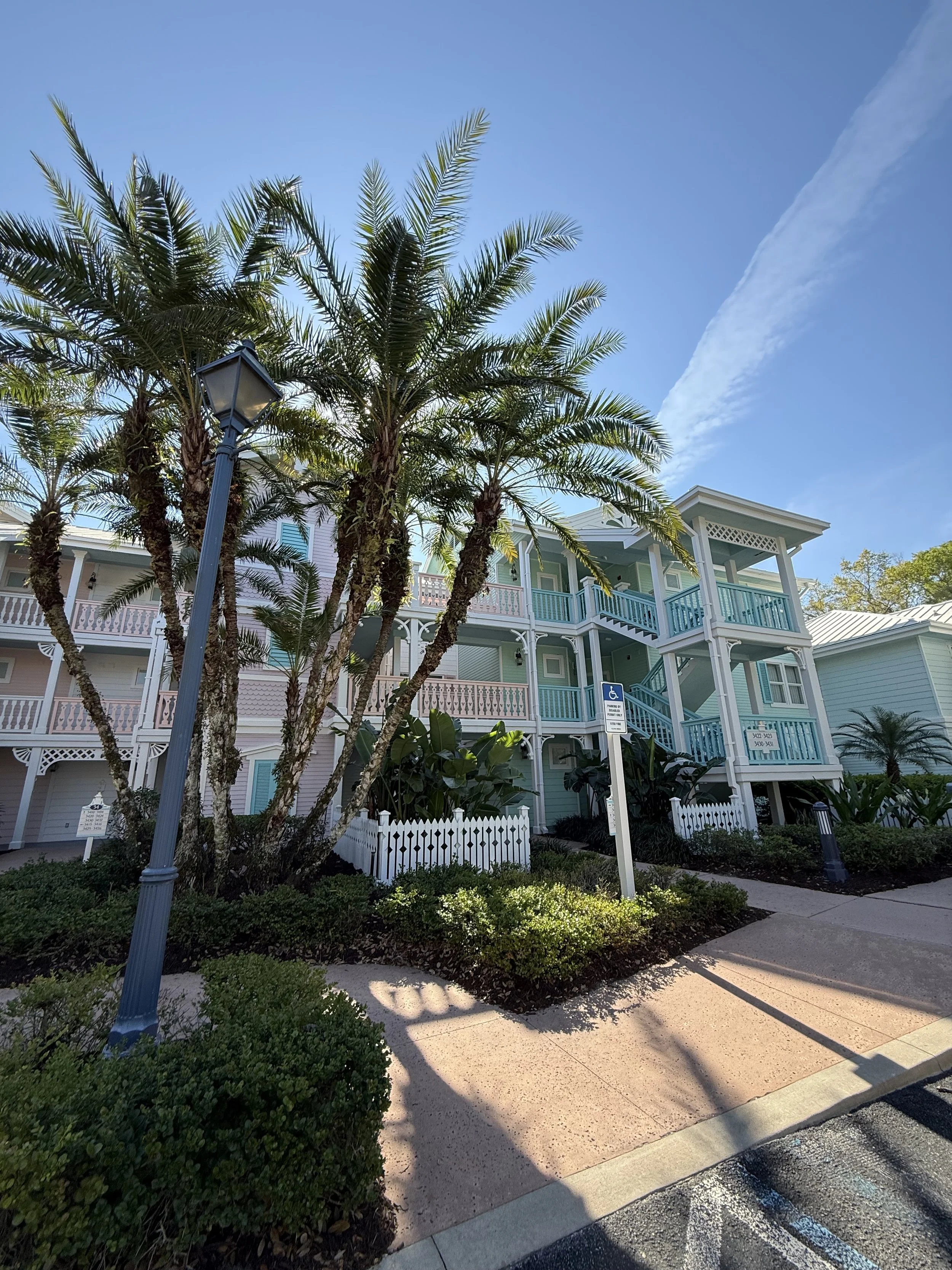 Colorful multi-story residential building with porches, surrounded by palm trees,, a street lamp, a handicapped parking sign, and well-maintained landscaping.