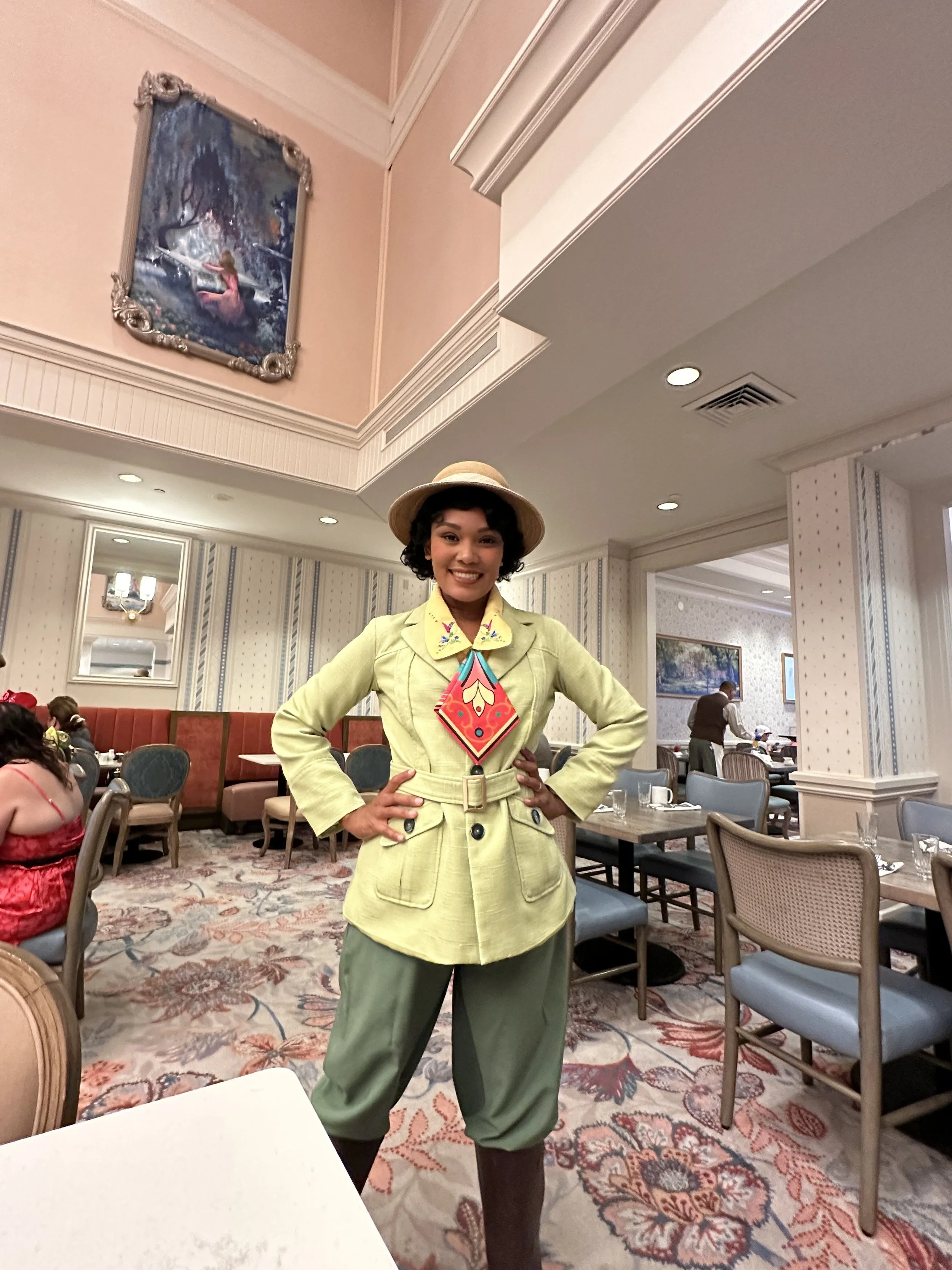 A woman in vintage outfit, hat, and colorful scarf standing in a restaurant with decorated ceiling and wallpaper, smiling at the camera.