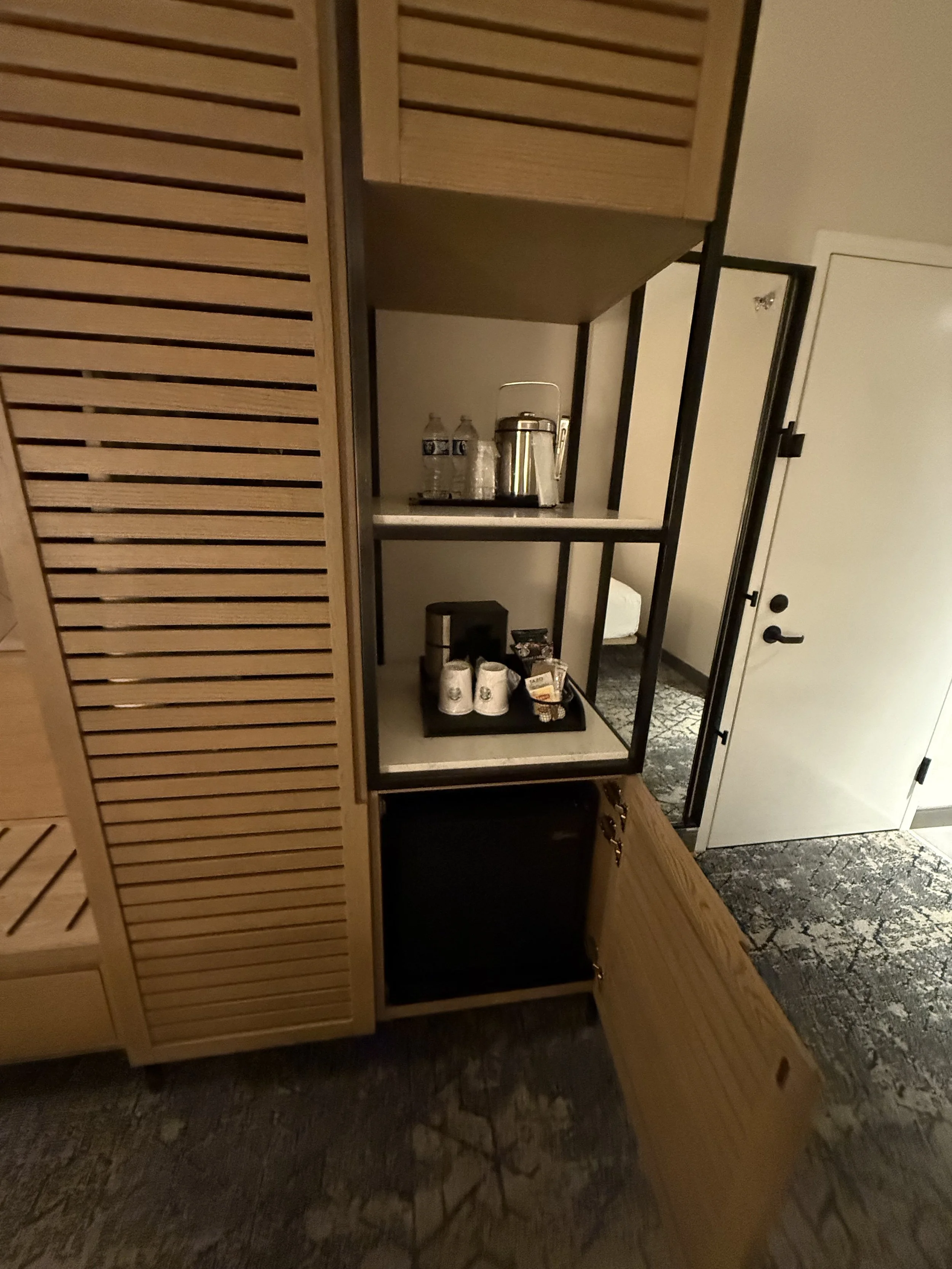 Coffee station inside a hotel room with coffee maker, cups, and bottled water on a black tray, and an open cabinet door revealing storage space below.