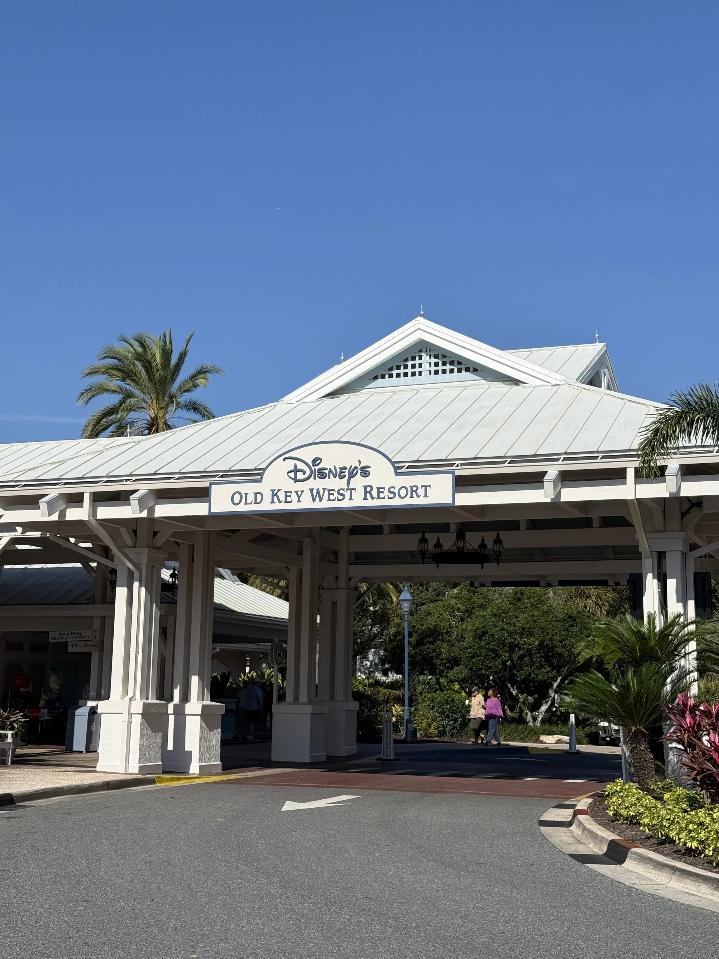 Entrance to Disney's Old Key West Resort with a white roof, sign, palm trees, and a clear blue sky.