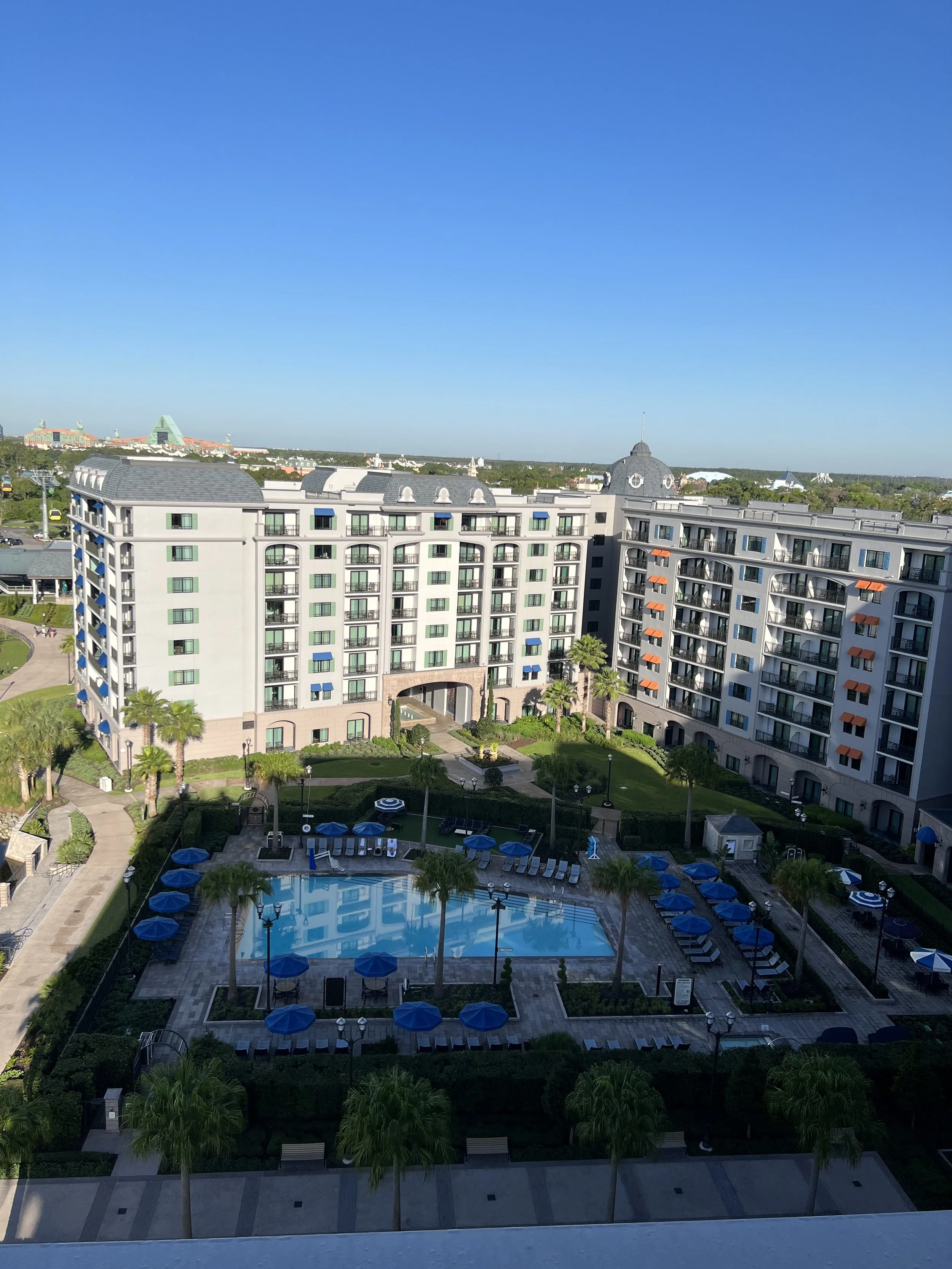 View of a residential complex with mid-rise buildings, a swimming pool with blue umbrellas, palm trees, and landscaped gardens under a clear blue sky.