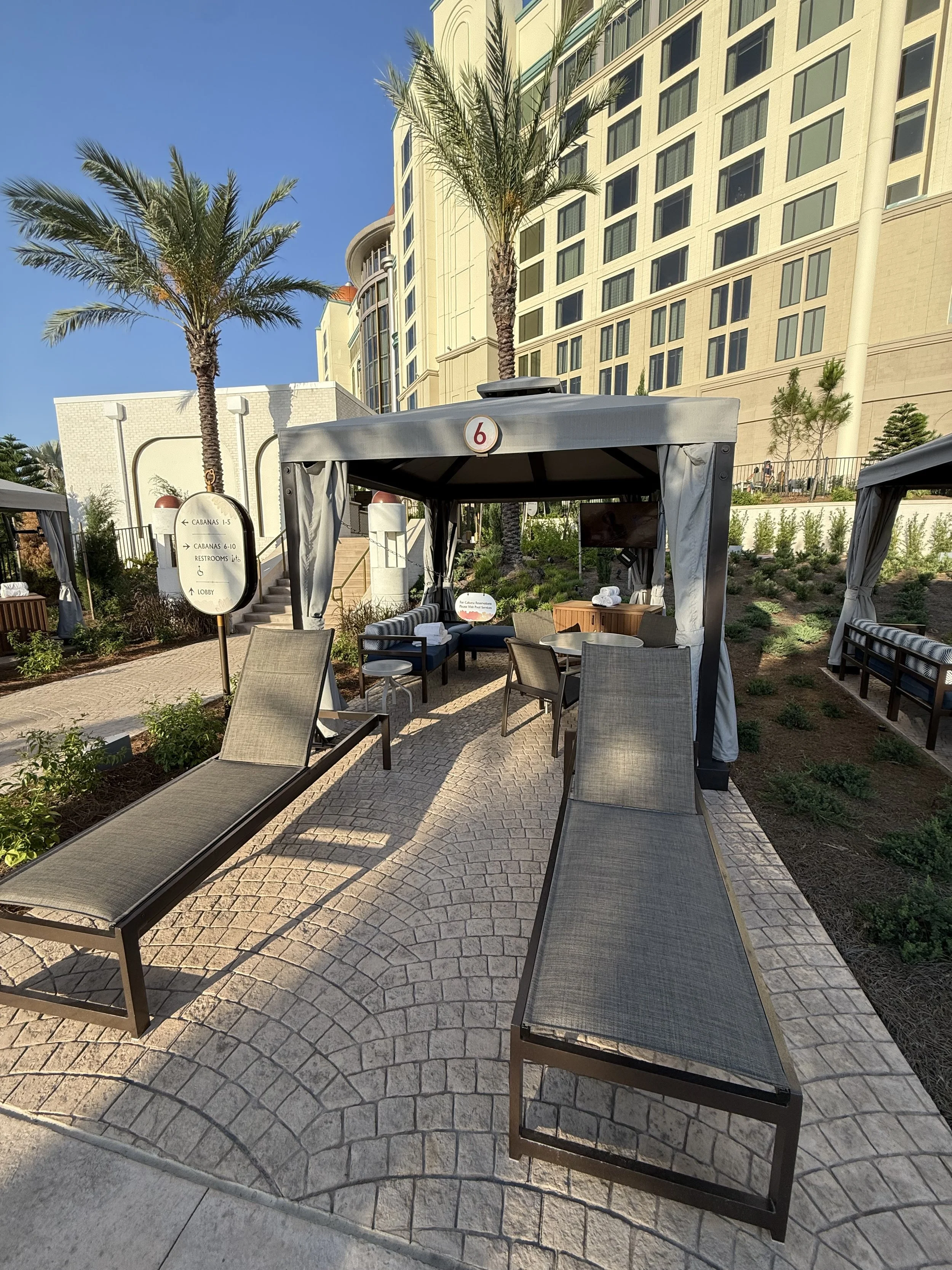 Outdoor resort lounge area with two beige lounge chairs, a cabana with seating and a TV, surrounded by palm trees and modern hotel building in the background.