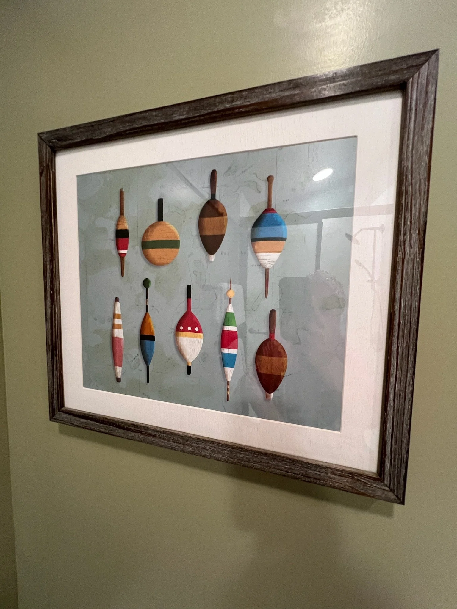 Framed wall art featuring ten colorful wooden fishing bobbers with various striped patterns hanging on a green wall.