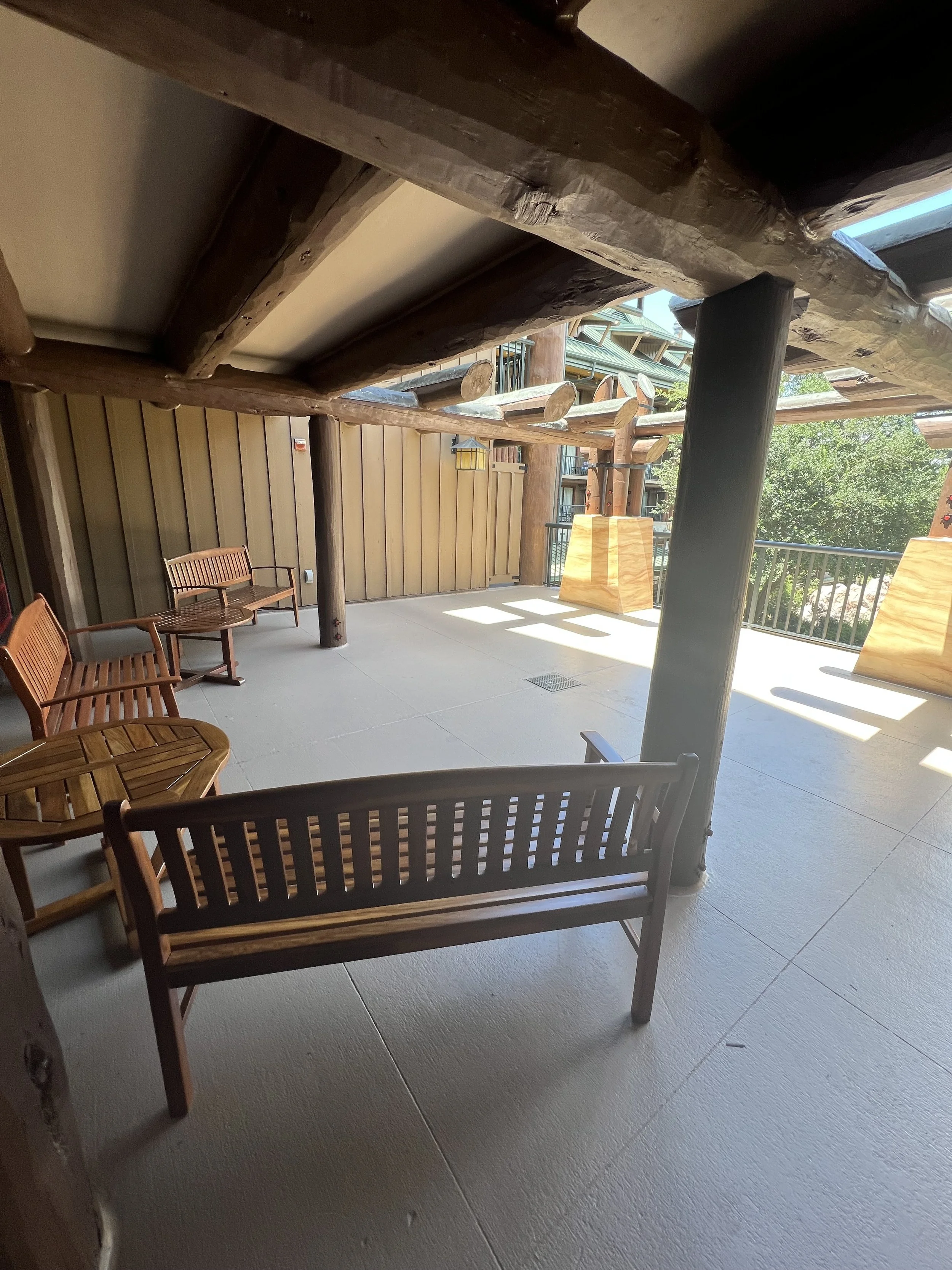 An outdoor patio area with wooden benches and chairs, a wooden fence, and large wooden decorative beams supporting the roof. Sunlight is shining onto the patio, creating shadows.