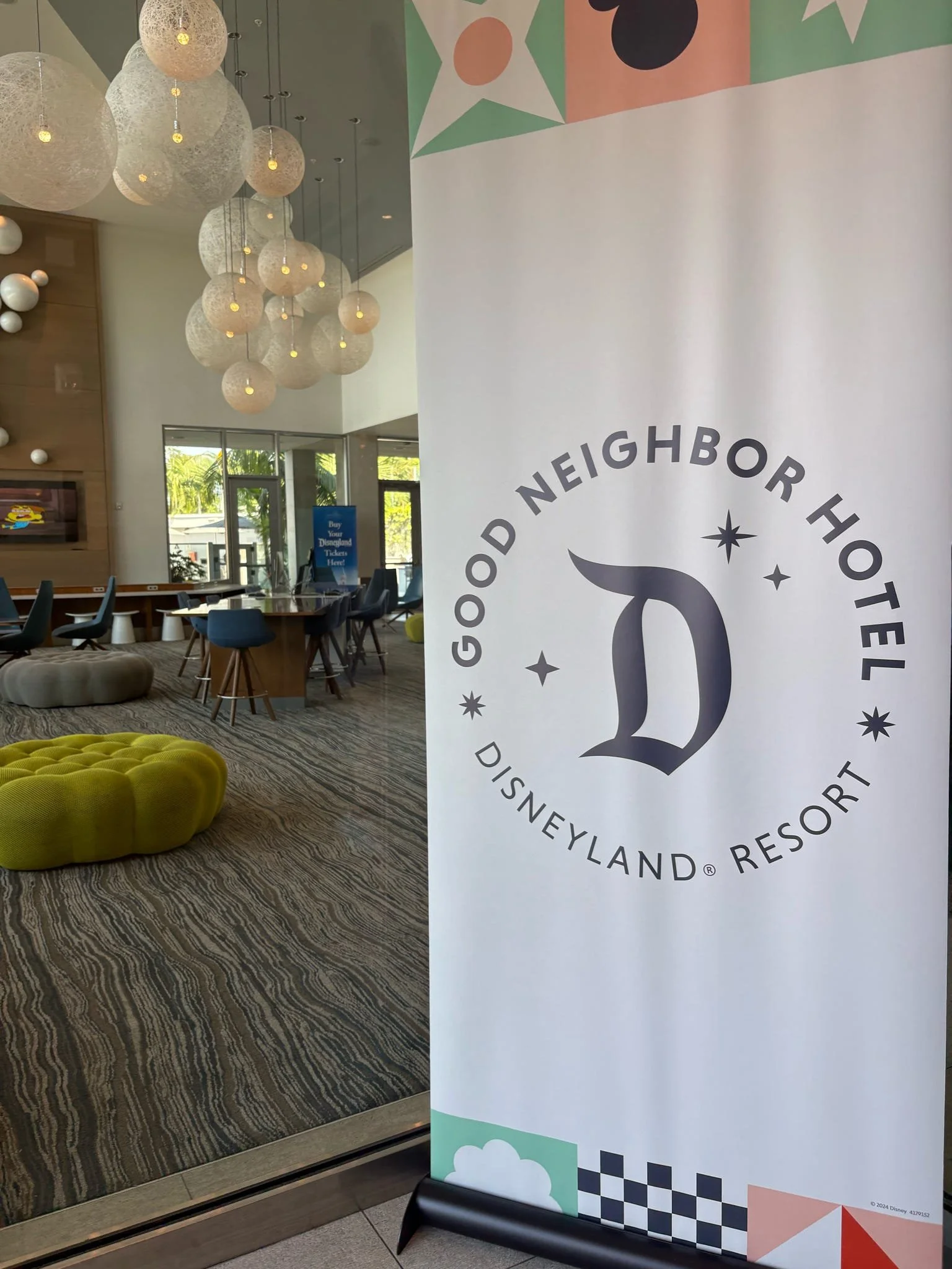 Interior of the Disneyland Resort hotel lobby with modern decor, a large banner displaying the hotel logo, hanging spherical light fixtures, and a seating area with colorful furniture.