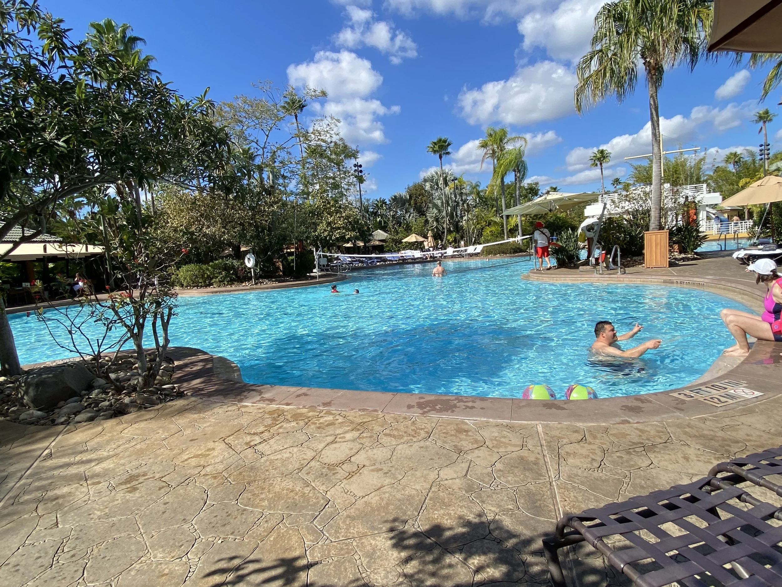 People enjoying a sunny day in and around a swimming pool at a resort, with palm trees, umbrellas, and lounge chairs visible.