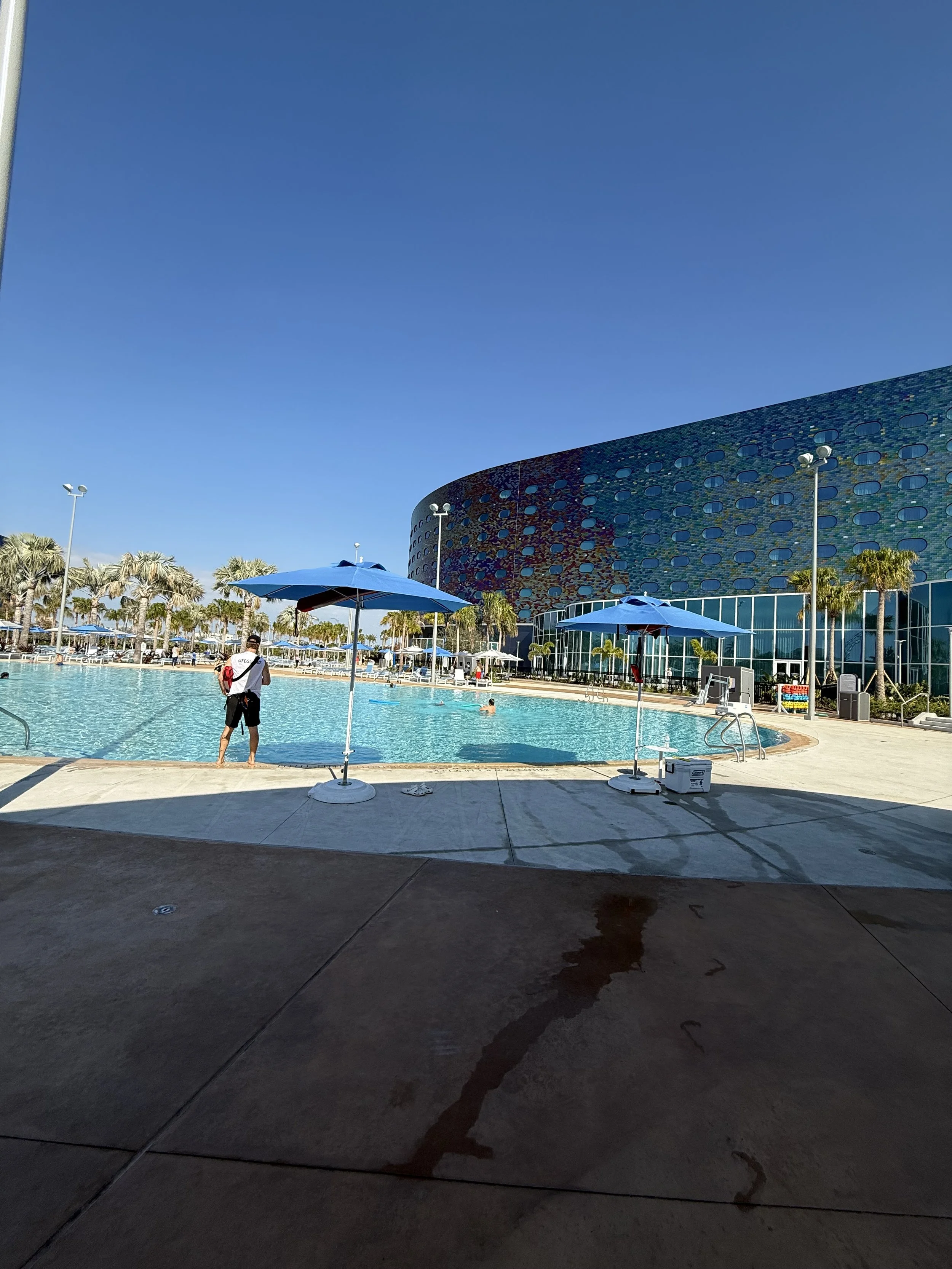 Outdoor swimming pool with blue umbrellas, a person standing by the pool, and a modern building with a rounded facade and circular windows in the background under a clear blue sky.