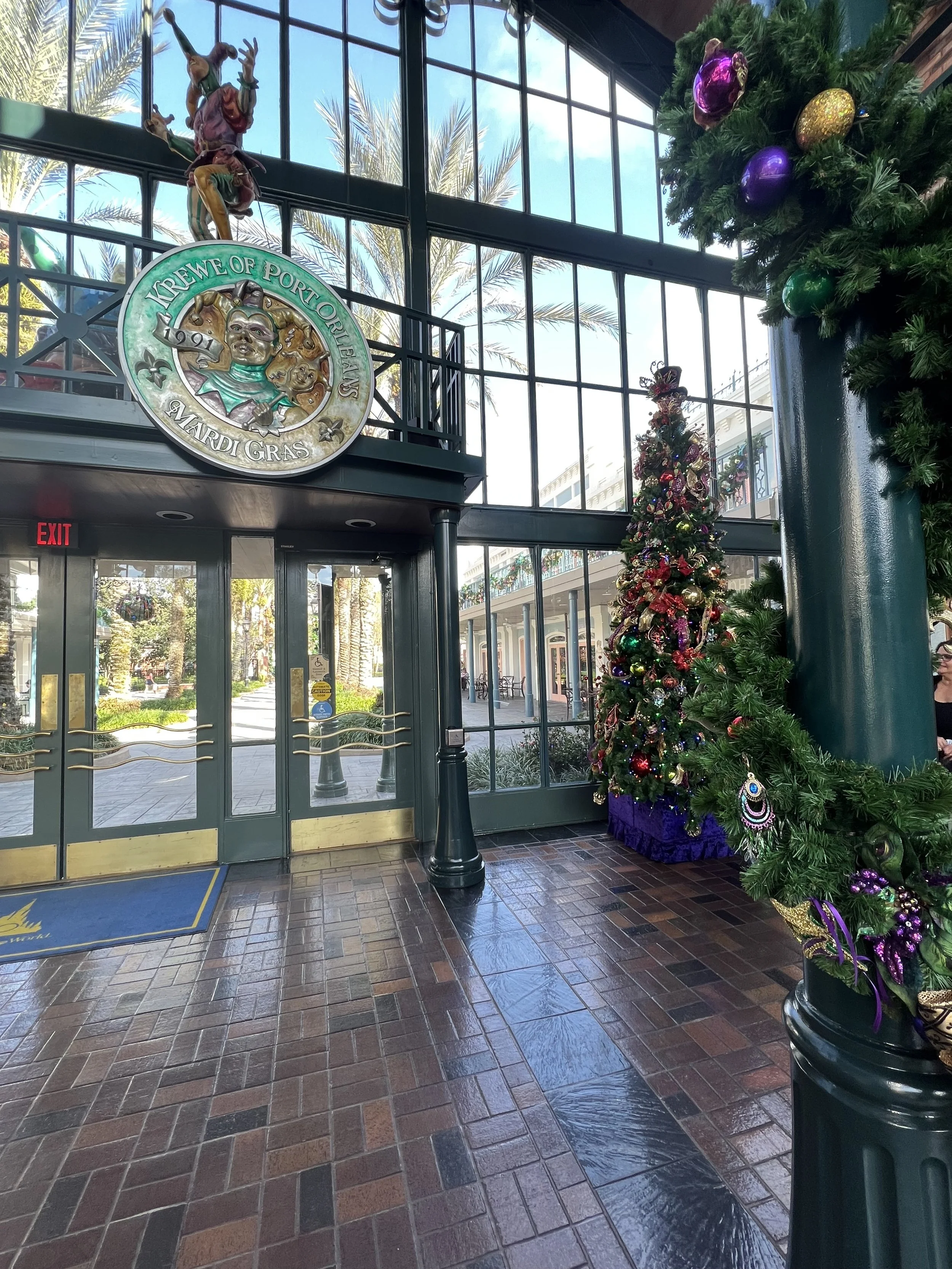 Decorated Christmas tree near entrance with glass doors, a Mardi Gras sign high above, and large windows showing palm trees outside.