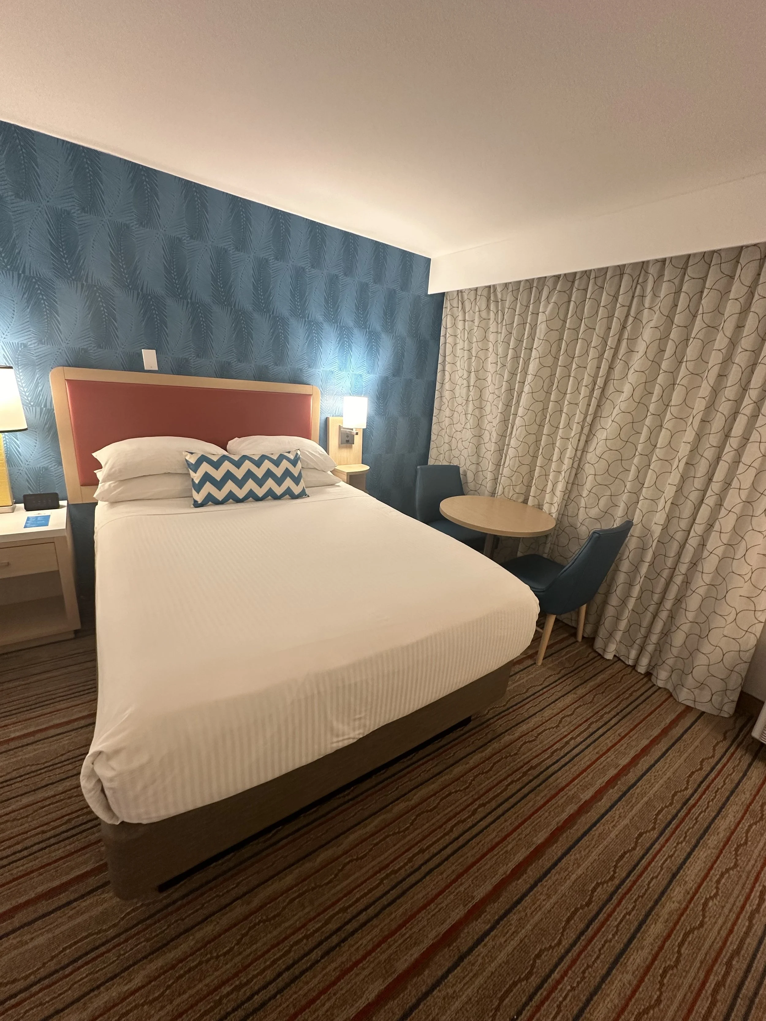 Hotel room with a bed, two bedside lamps, a blue accent wall behind the bed, a pillow with a chevron pattern, a small round table, and two blue chairs near a window with patterned curtains.