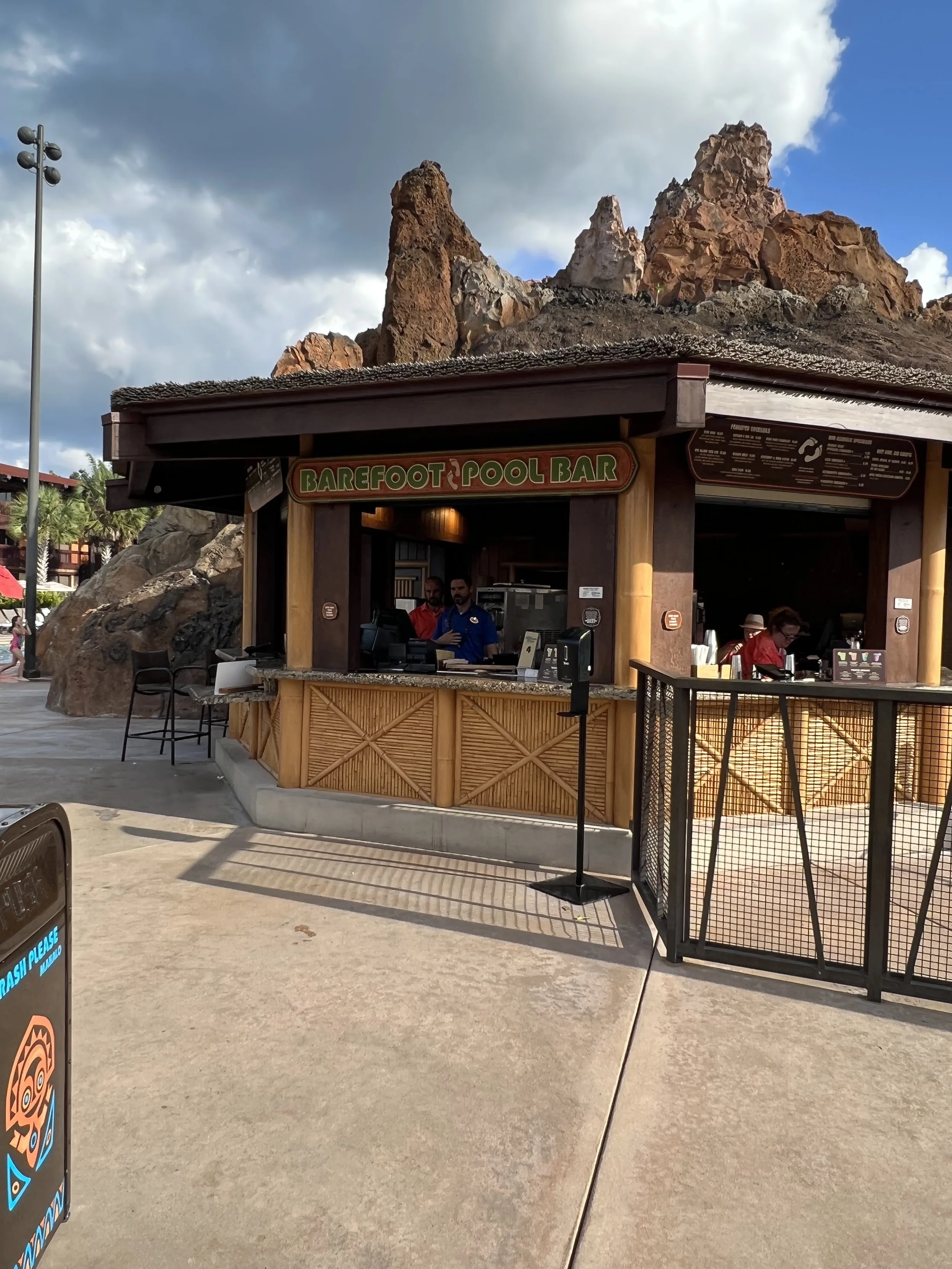 A small outdoor pool bar called 'Barefoot Pool Bar' with a wooden structure, situated against a backdrop of large rocks or mountains. There are people working behind the counter, and the sky is partly cloudy.