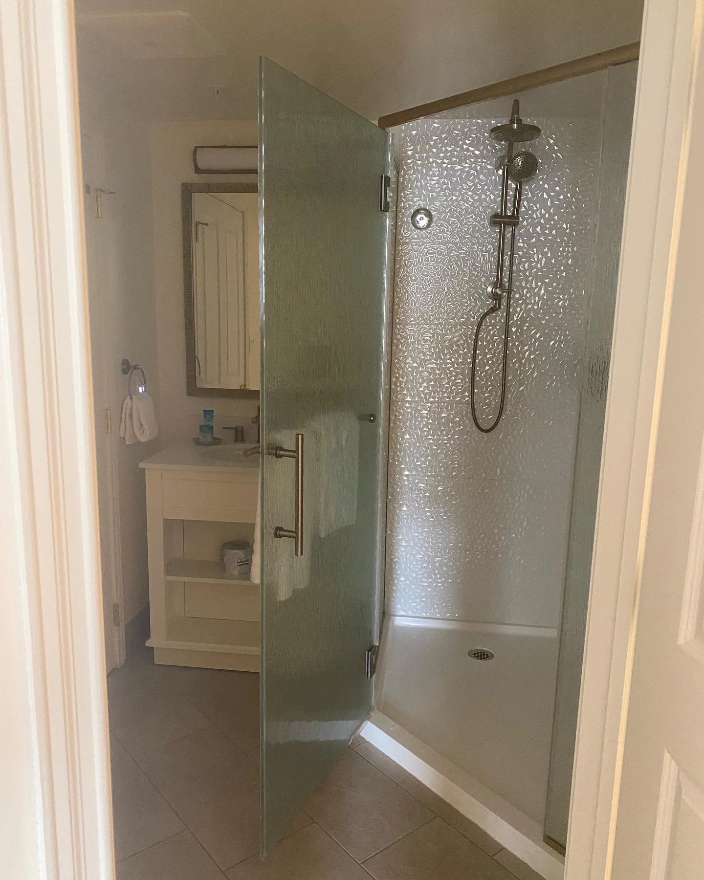 A bathroom shower with textured glass walls and a stainless steel showerhead. There is a door that opens inward. Behind the shower, part of a white vanity with a mirror and towel. The floor is tiled in beige.