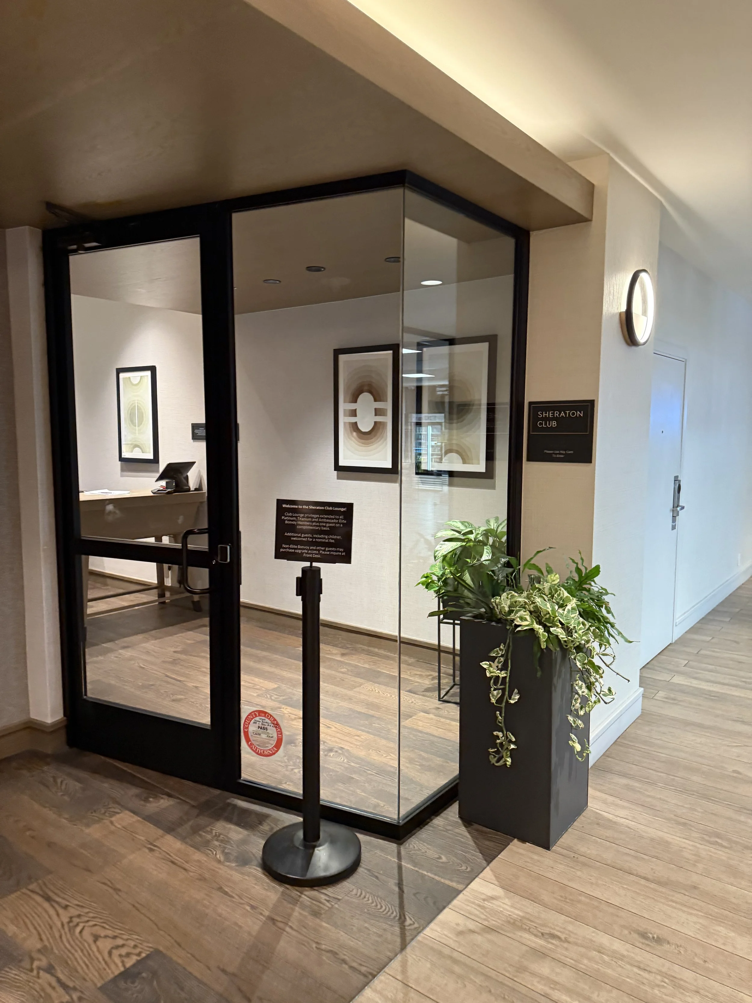 Entrance to the Sheraton Club Lounge with glass walls, a sign on the wall, and a potted plant outside.