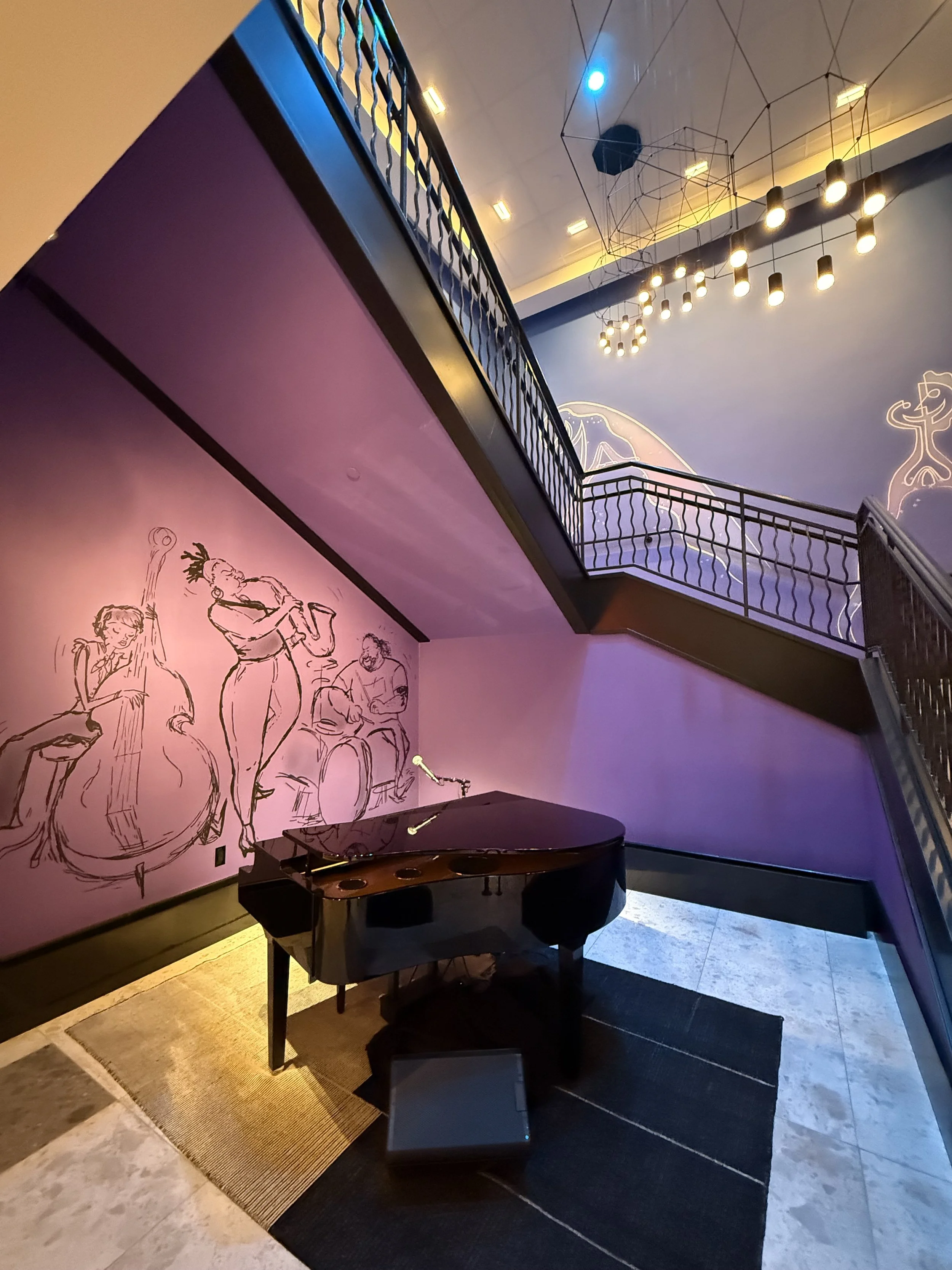 Interior of a music lounge featuring a black grand piano and a mural of jazz musicians on the purple wall, with a staircase, modern lighting, and decorative ceiling fixtures.
