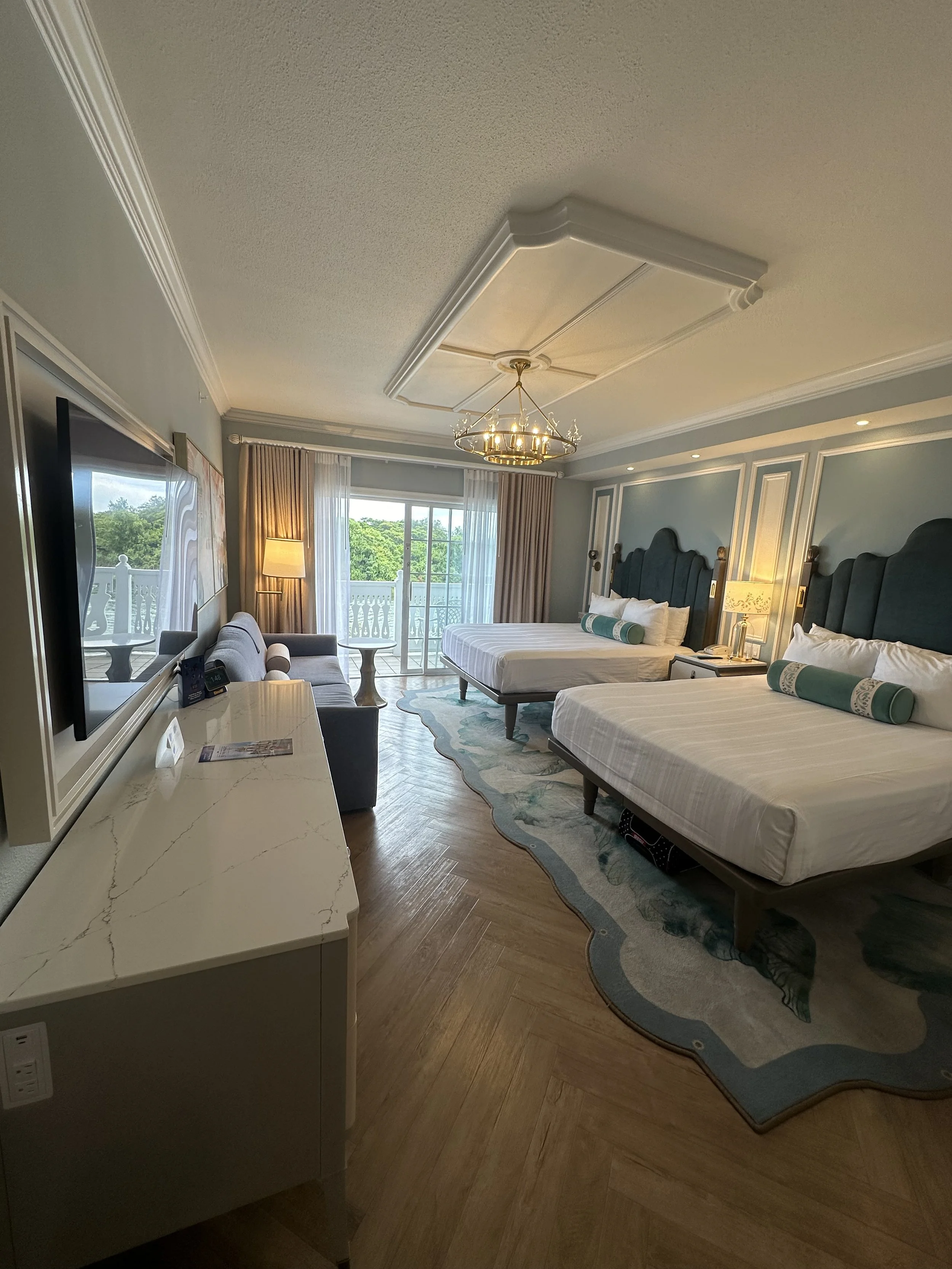 A hotel room with two queen beds, a sliding glass door leading to a balcony, a sitting area with a sofa, a large flat-screen TV, and decorative lighting fixtures.