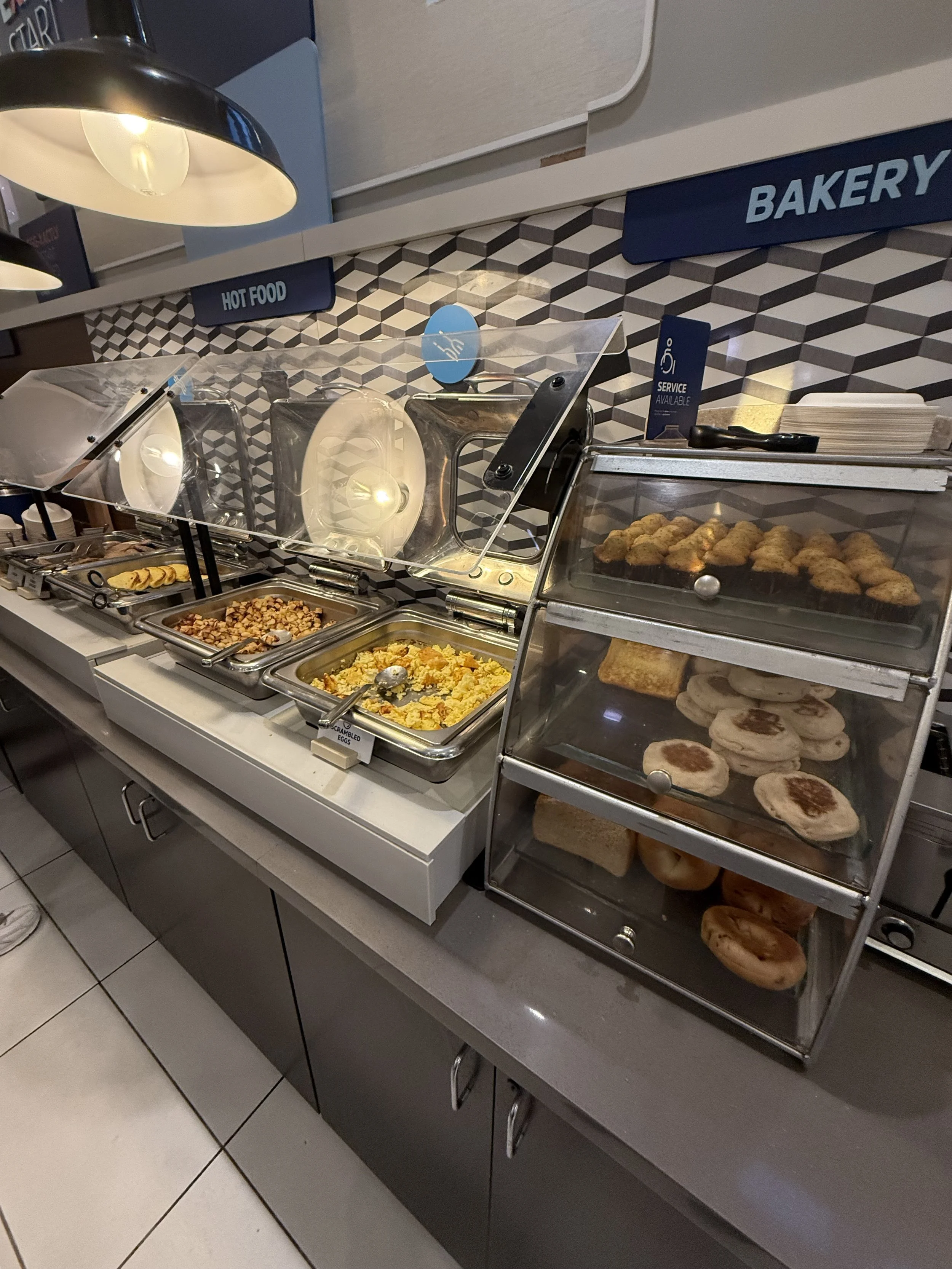 A breakfast buffet with hot food options and baked goods including scrambled eggs, fried potatoes, muffins, and bagels.