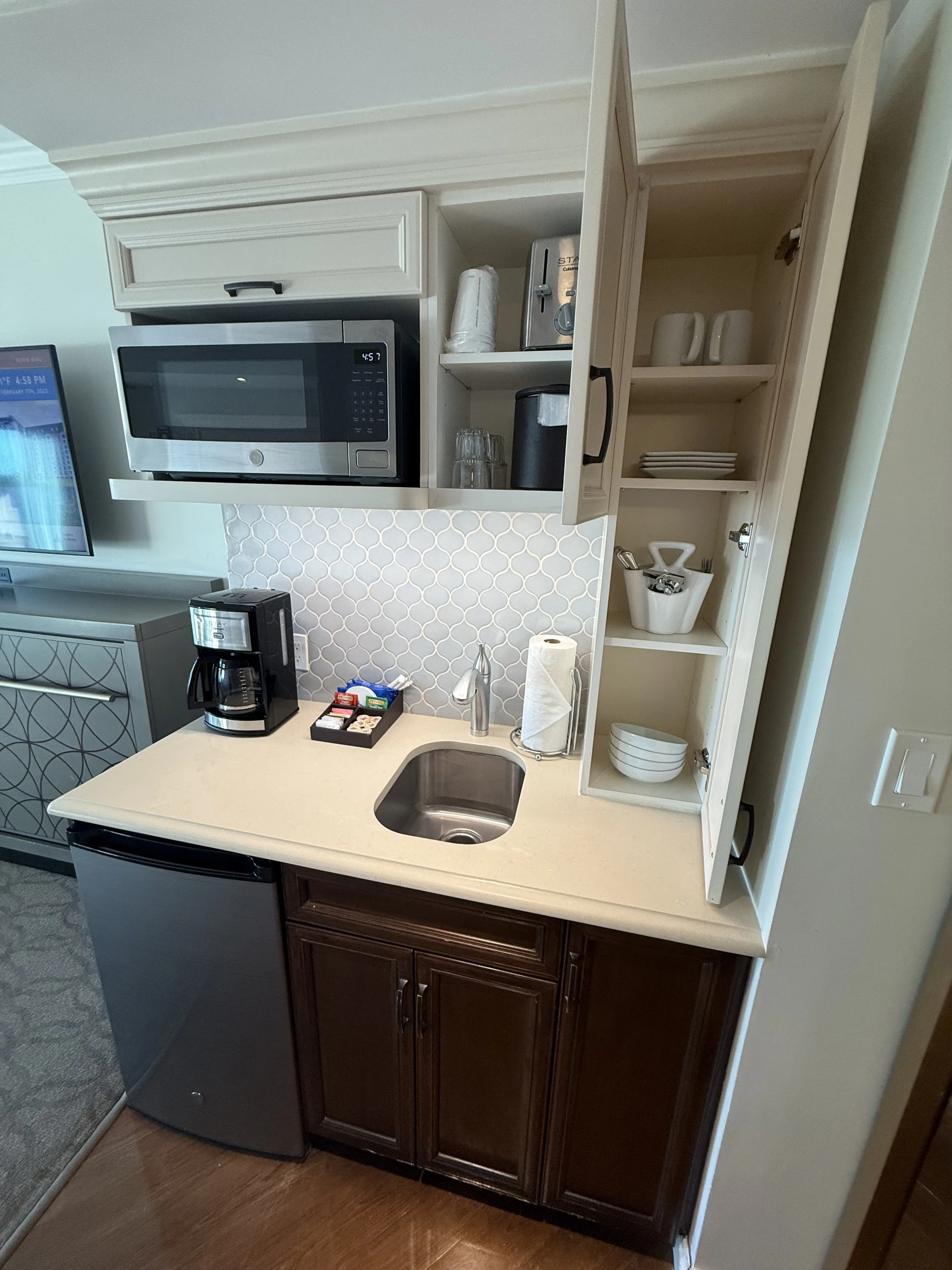 Miniature kitchenette with a small sink, coffee maker, microwave, small refrigerator, wooden cabinets, and open shelves with dishes and cups.