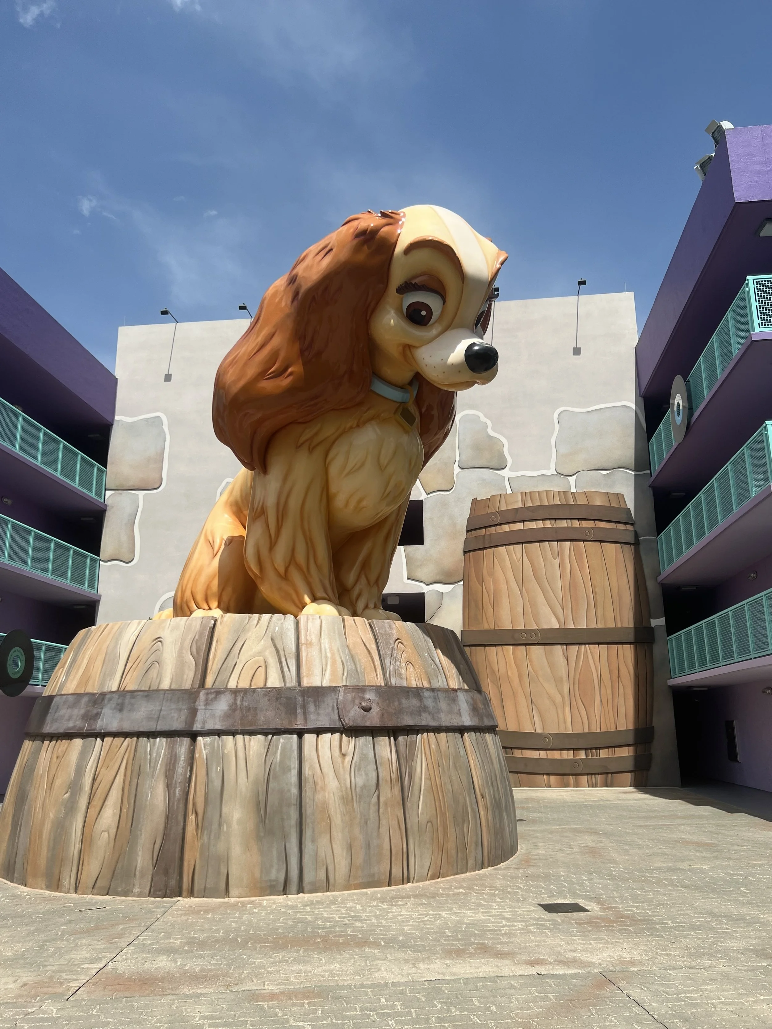 Large statue of Lady from Lady and the Tramp sitting on a wooden barrel in an outdoor area with purple apartment buildings and a clear blue sky.