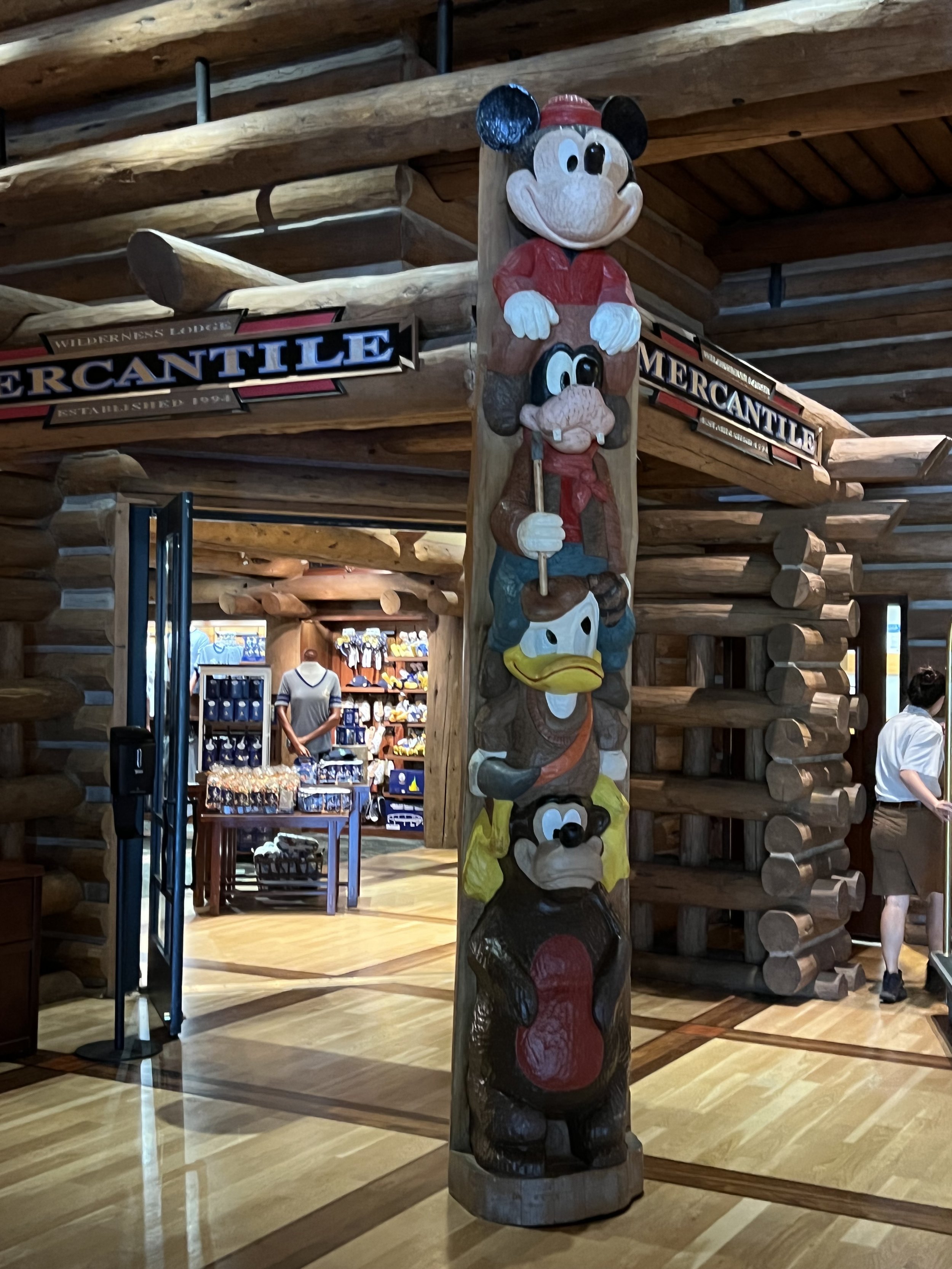 Colorful wooden totem pole featuring Disney characters Mickey Mouse, Goofy, Donald Duck, and Pluto inside a log cabin-style store.