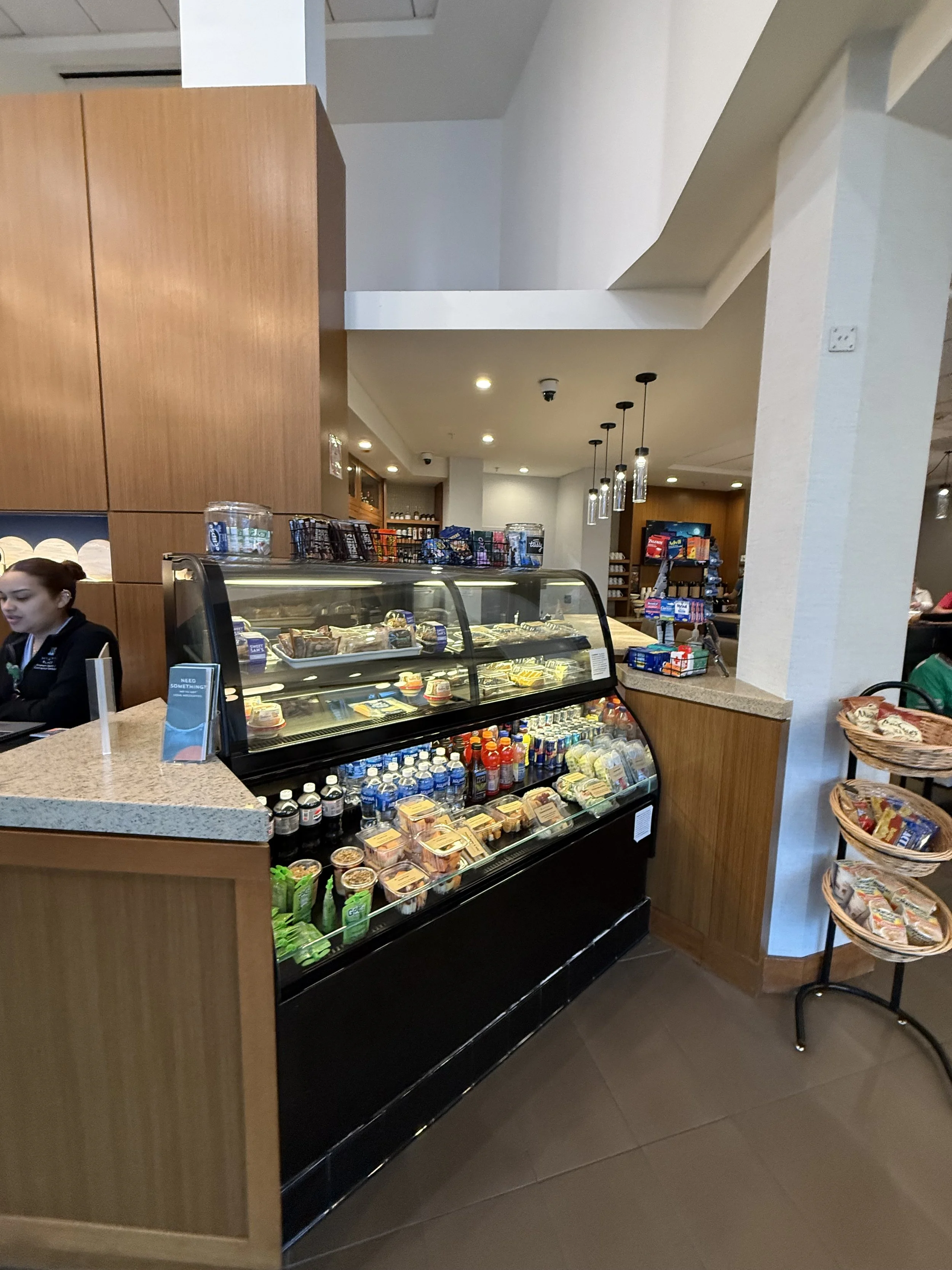 Inside a cafe showing a food and beverage display case with sandwiches, snacks, and drinks, next to a counter with a staff member and a snack stand with chips.
