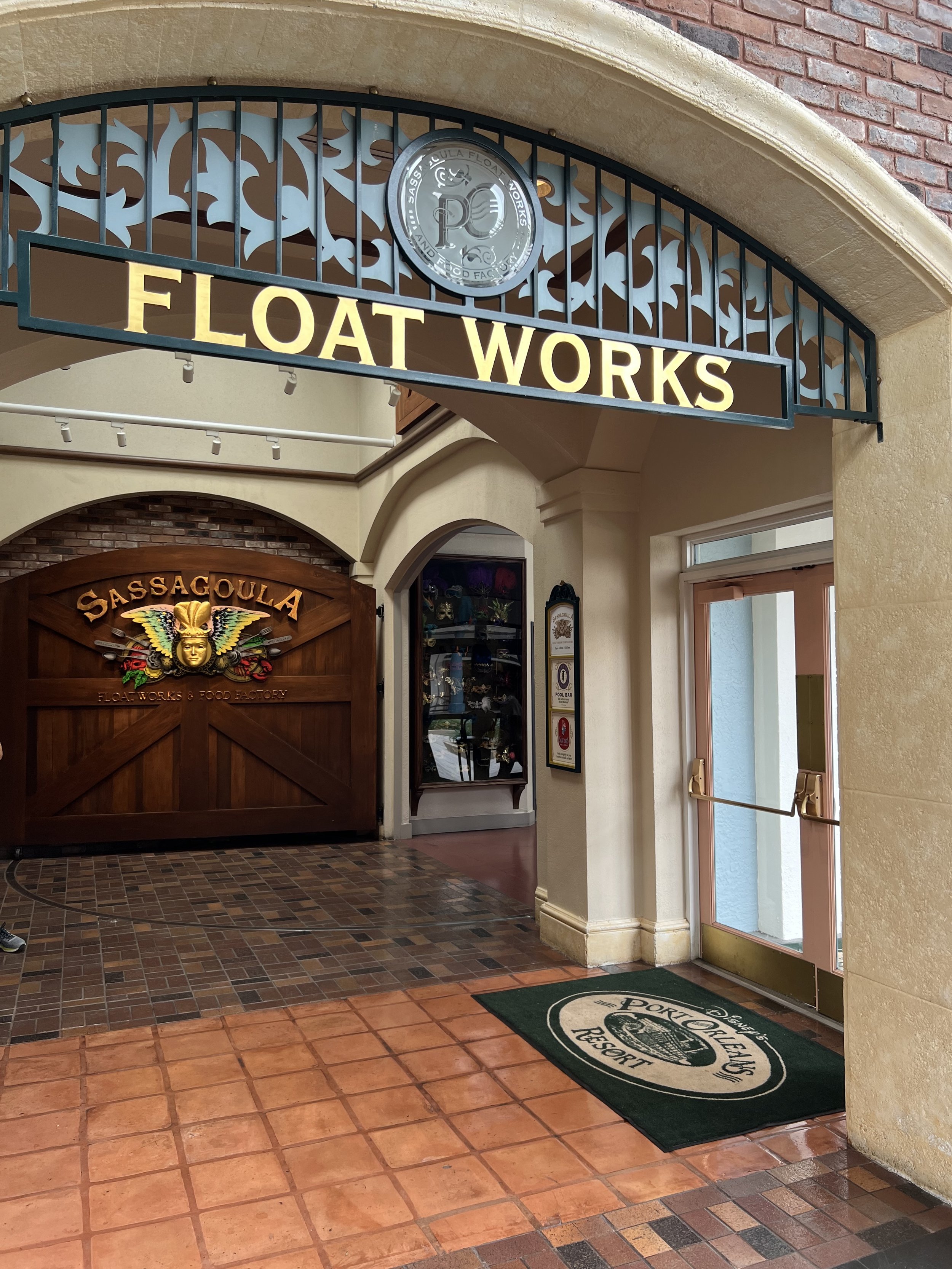 Entrance to the Sassa Gula Float Works & Food Factory, with a wooden gate featuring a colorful mask and wings, and a green port Orleans Resort mat at the door.