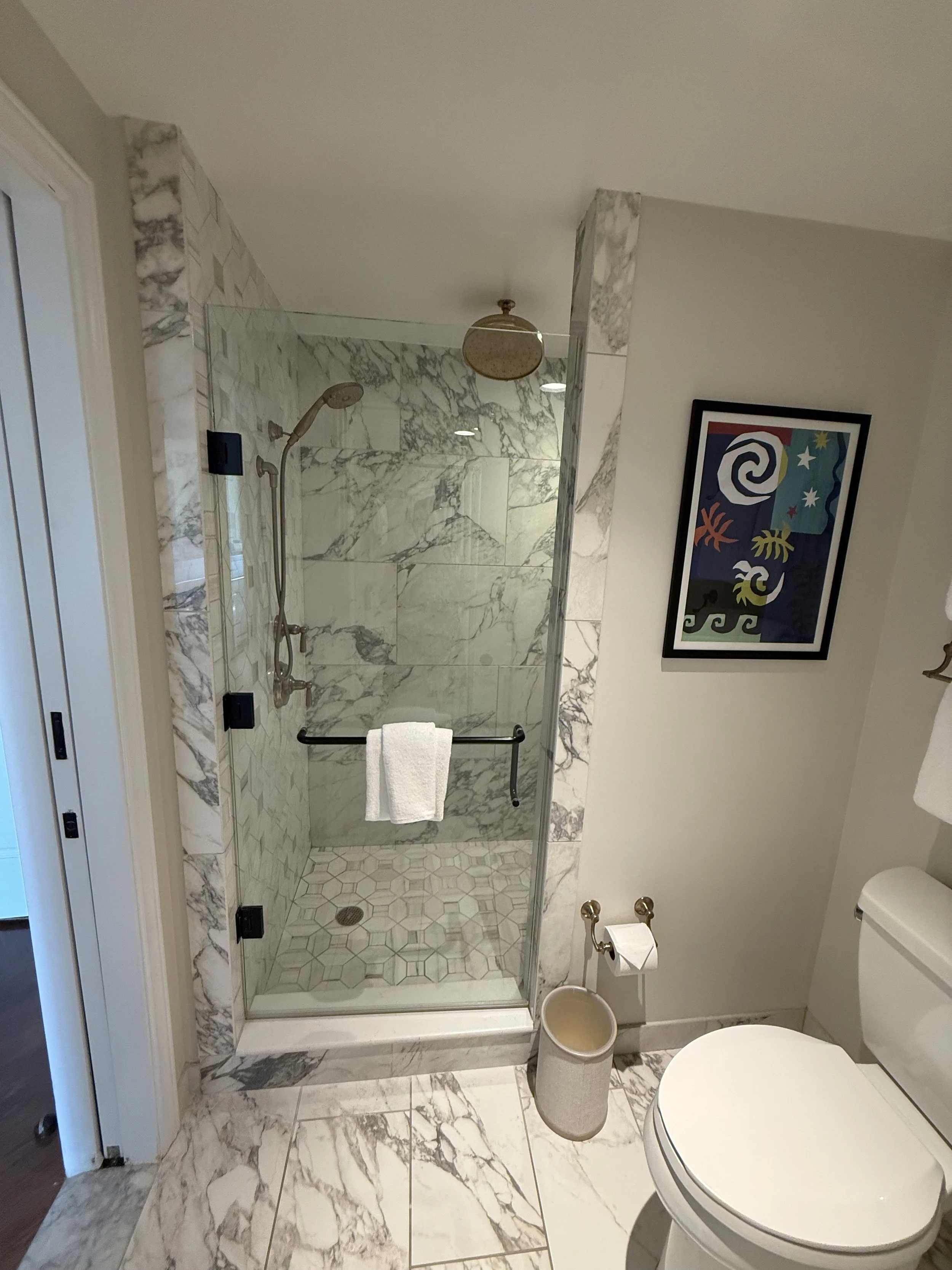 Bathroom with a marble shower, framed colorful abstract art, a toilet, and a trash bin.