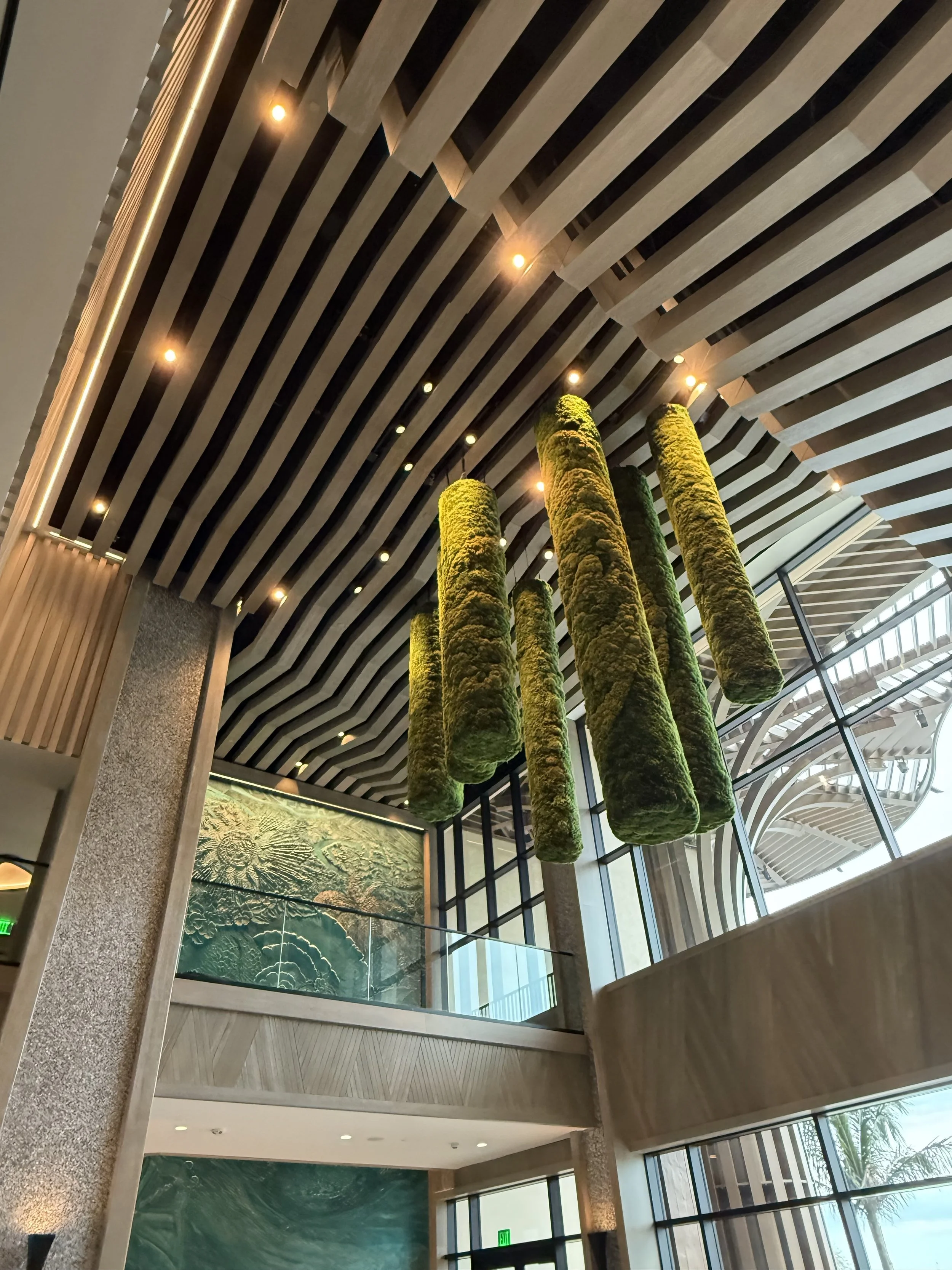 Interior of a modern building with a high ceiling featuring wooden slats and small recessed lights. Large green moss-like sculptures hang from the ceiling, and there are large windows letting in natural light.